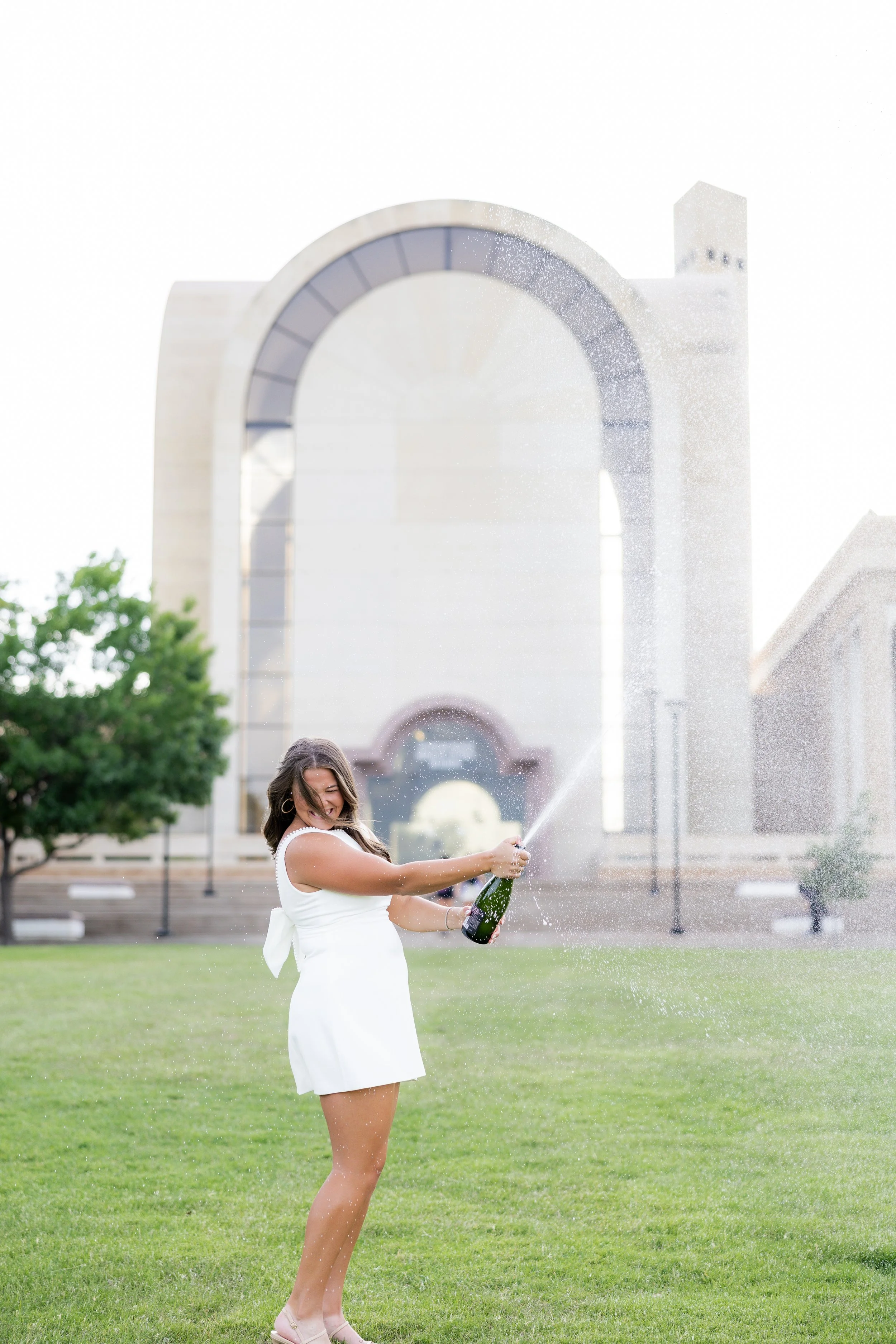 A woman in a white dress celebrates with a bottle of champagne and a champagne spray around her during graduation photography session.