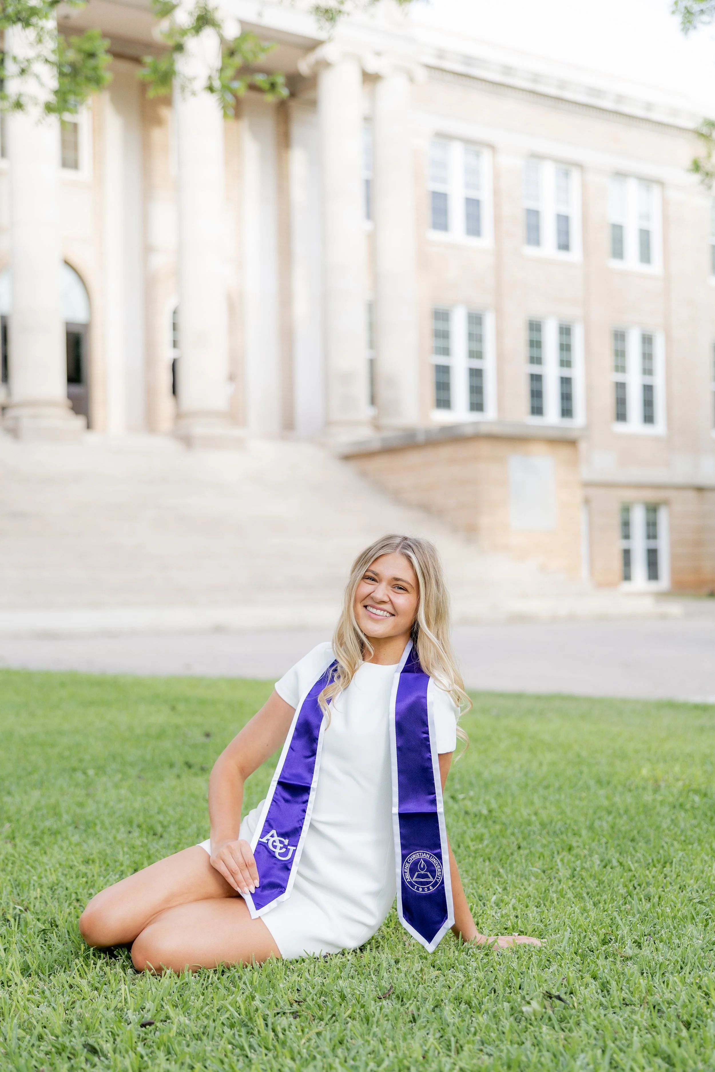 College graduate in a white dress and purple graduation stole posing on campus.