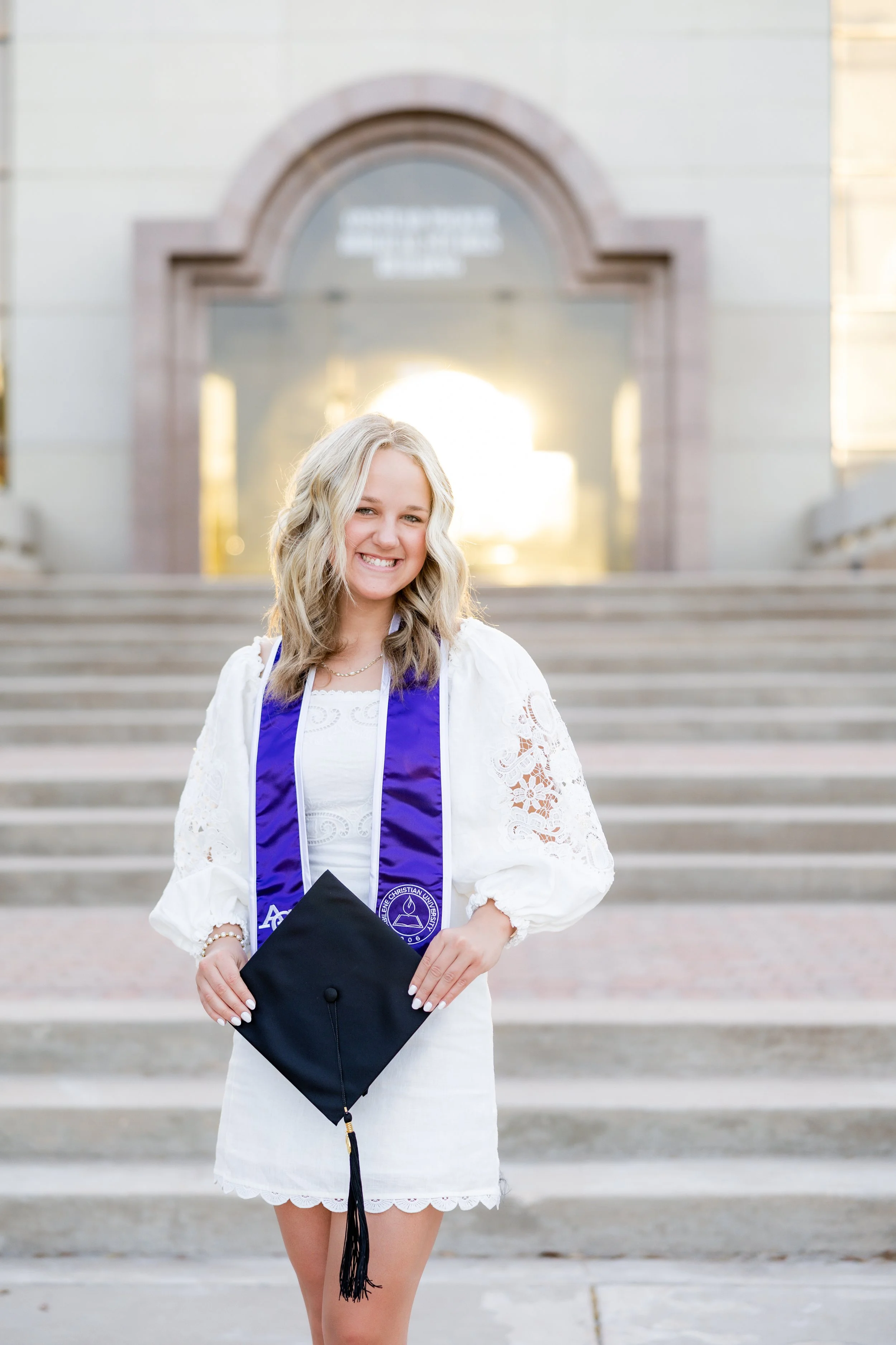 College graduate in a white dress and graduation stole holding her cap during an outdoor graduation session.