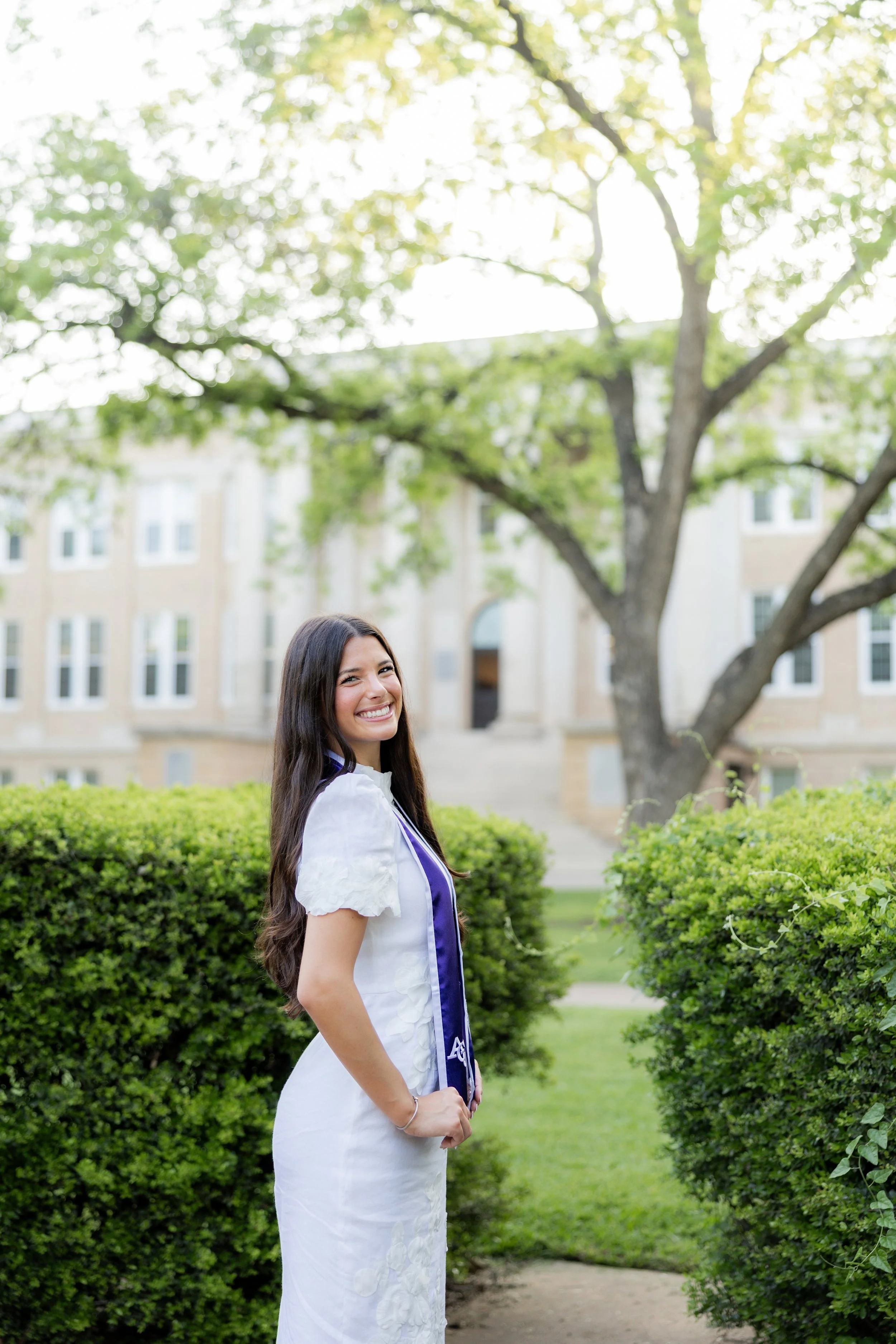 College graduate in a white dress and graduation stole posing outdoors with trees.