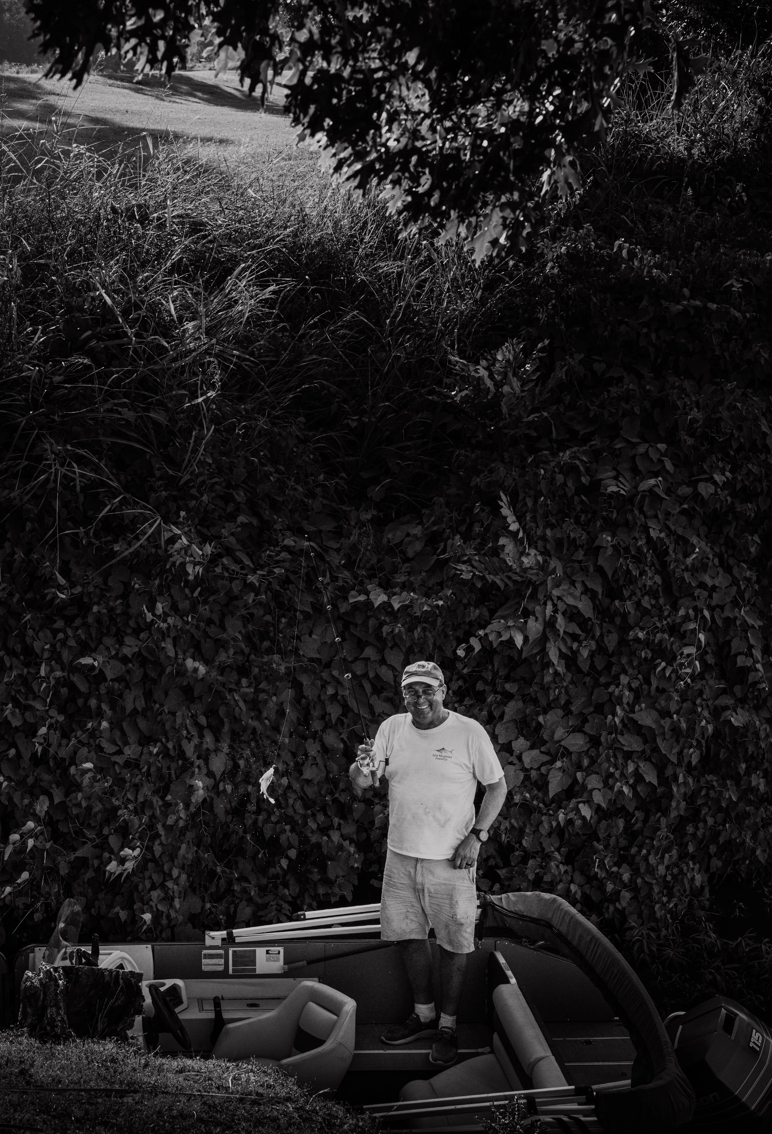 A man standing in a boat filled with fishing gear, smiling, wearing a cap and casual clothes, outdoors near a lush, dense hedge.