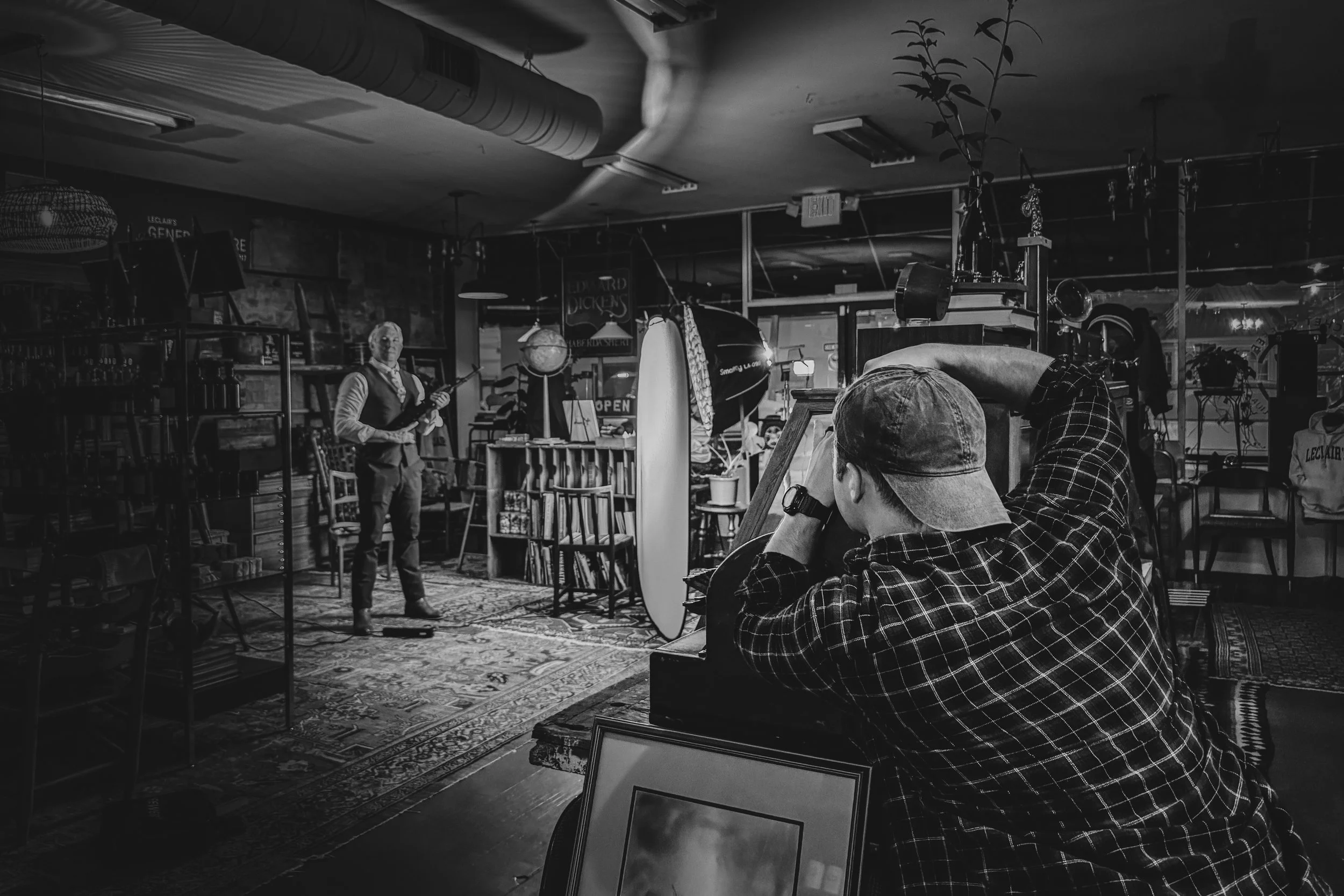 A black and white photo of a person in a plaid shirt taking a photograph of a man holding a guitar in a vintage-style store or cafe with sculptures and shelves visible in the background.