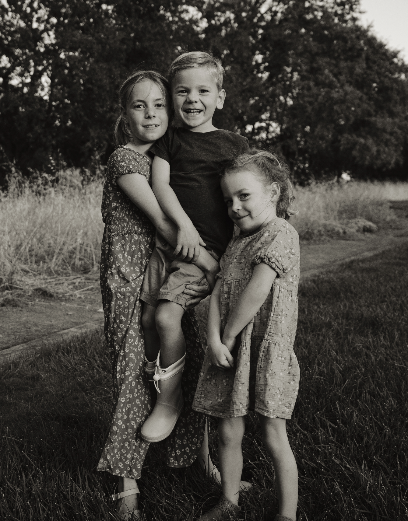 Three children, two girls and a boy, standing outdoors on grass with trees in background. The boy is being held by the girls, smiling and looking at the camera. One girl has her arm around the boy's shoulders, and the other girl is standing beside th