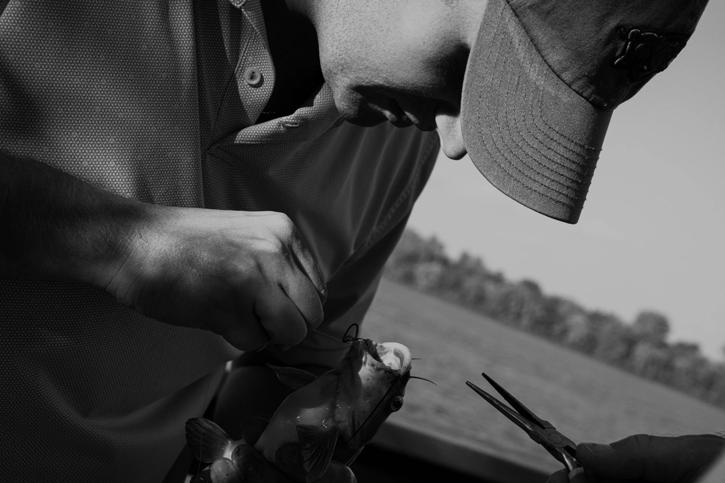 A person wearing a baseball cap and patterned shirt holding fishing gear outdoors, with landscape in the background.