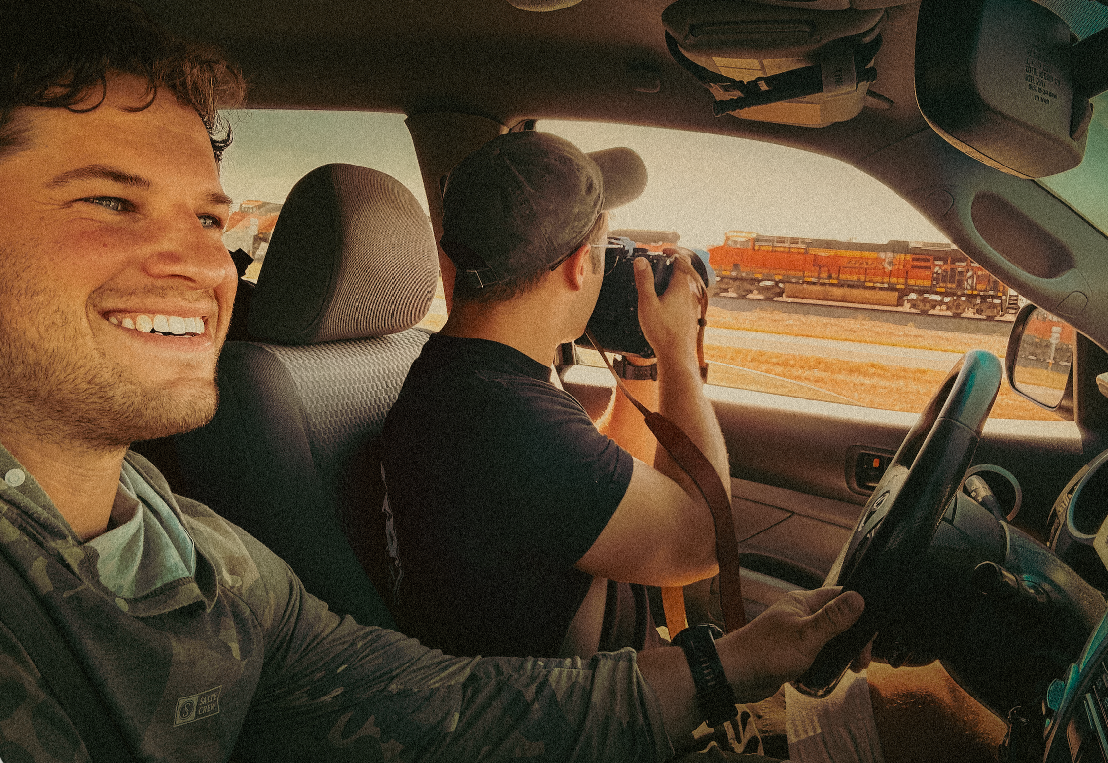 Two men inside a vehicle, one smiling at the camera and the other wearing a camouflage cap, holding a camera, with a train visible outside through the window.