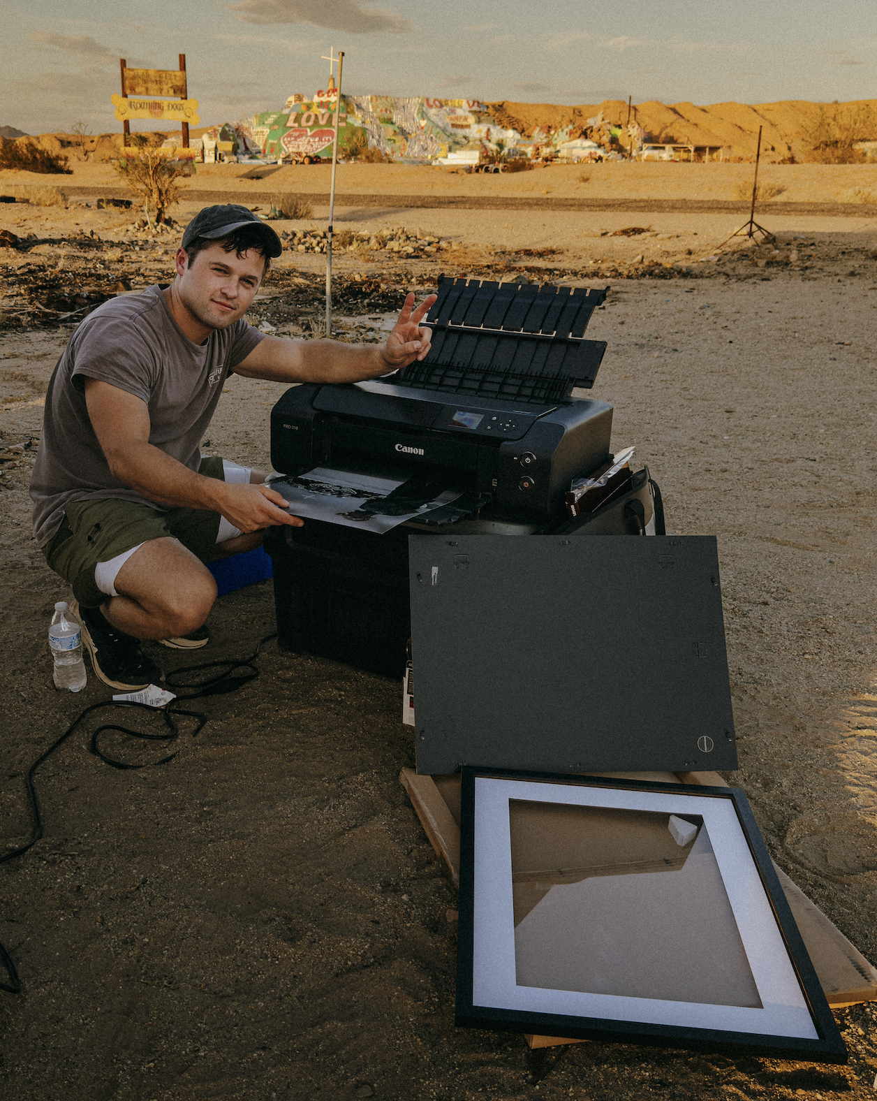 A young man outdoors using a Canon printer, making a peace sign, with photo frames and artwork around him, in a desert-like area with graffiti on distant hills.