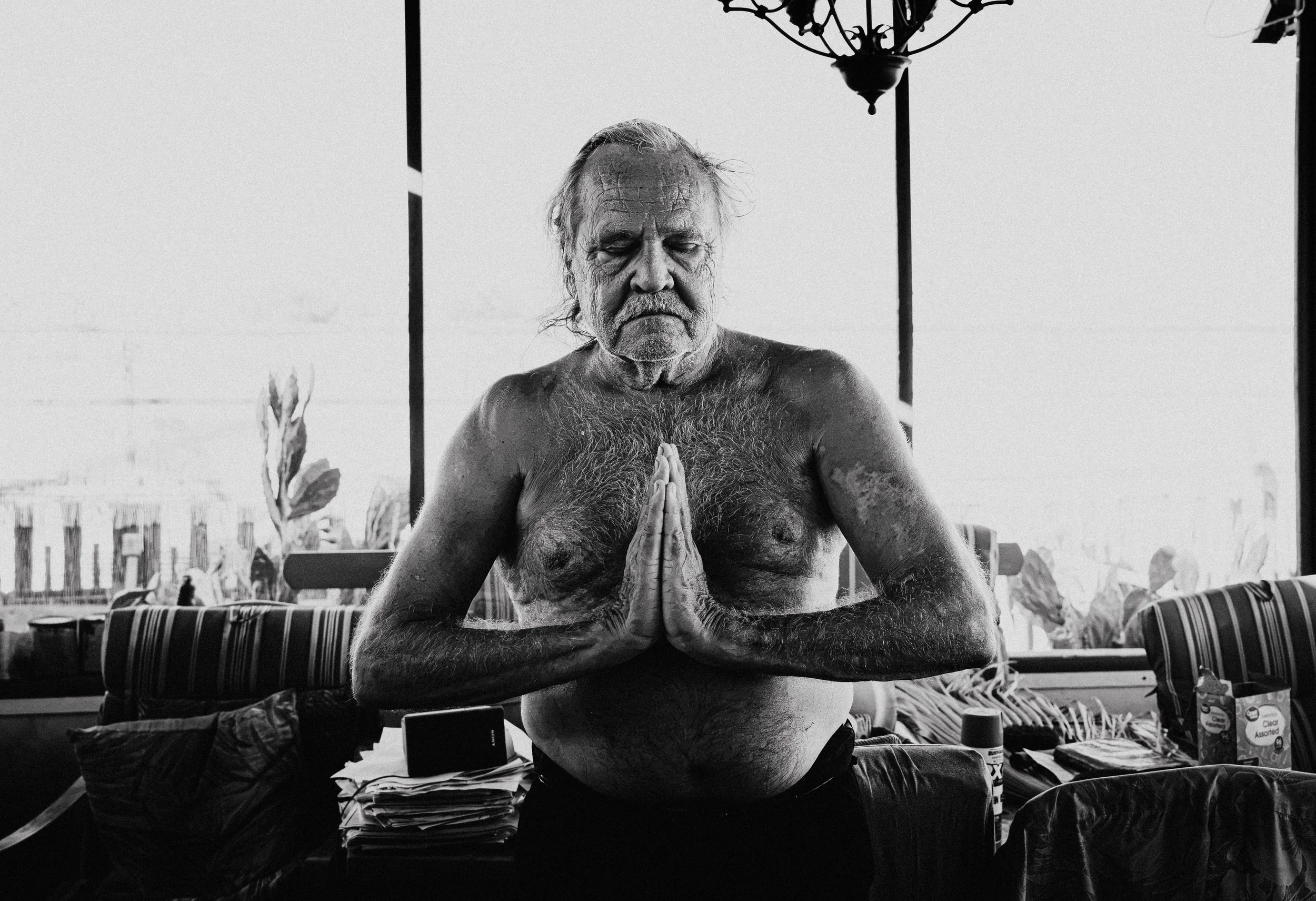 An elderly man with a beard and long hair practicing yoga outdoors, sitting shirtless with hands pressed together in a prayer position, eyes closed, under a hanging chandelier, with items on a table behind him.