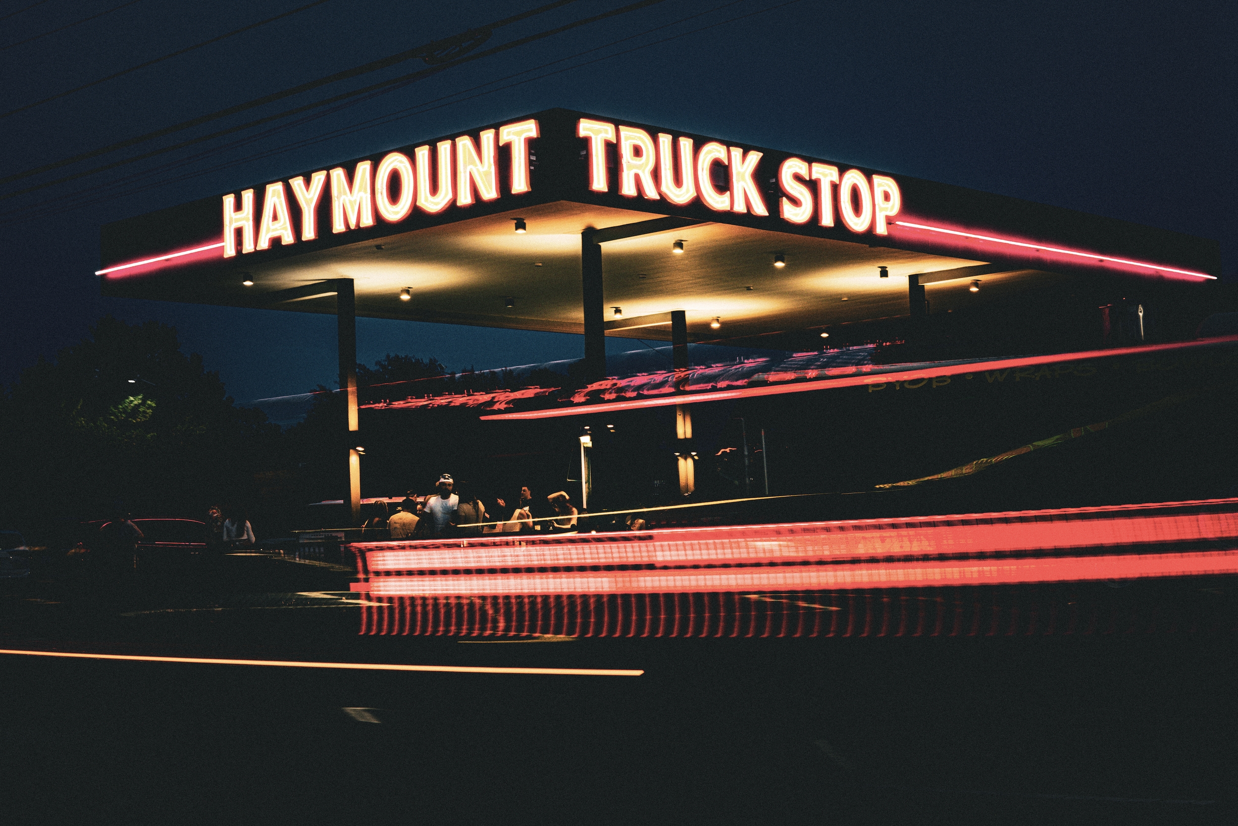 Nighttime scene of the Haymount Truck Stop with bright neon signs and people gathering under the canopy.