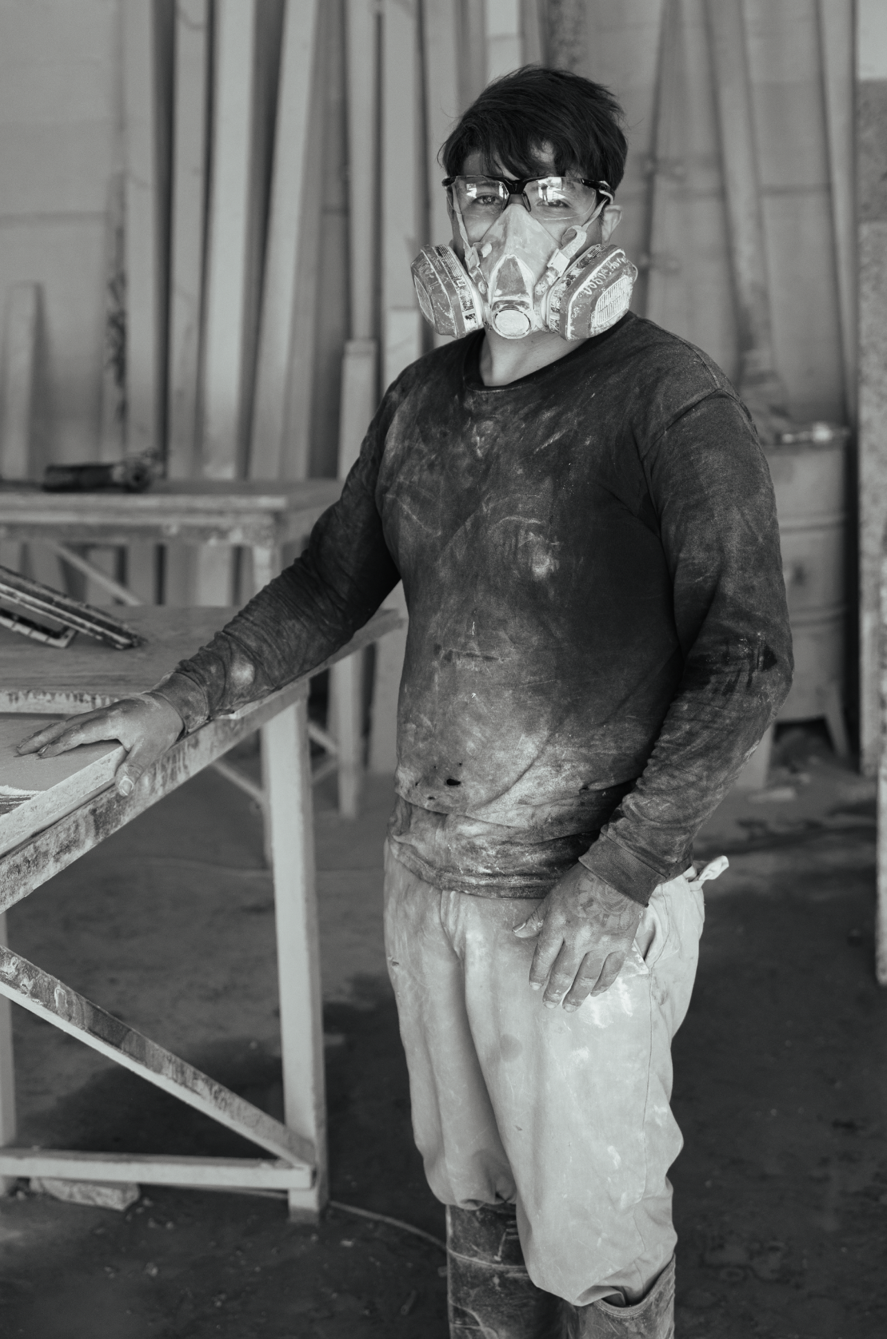 A construction worker wearing safety glasses, a dust mask, and work clothes covered in dust and dirt standing in a workshop with wood and construction tools.