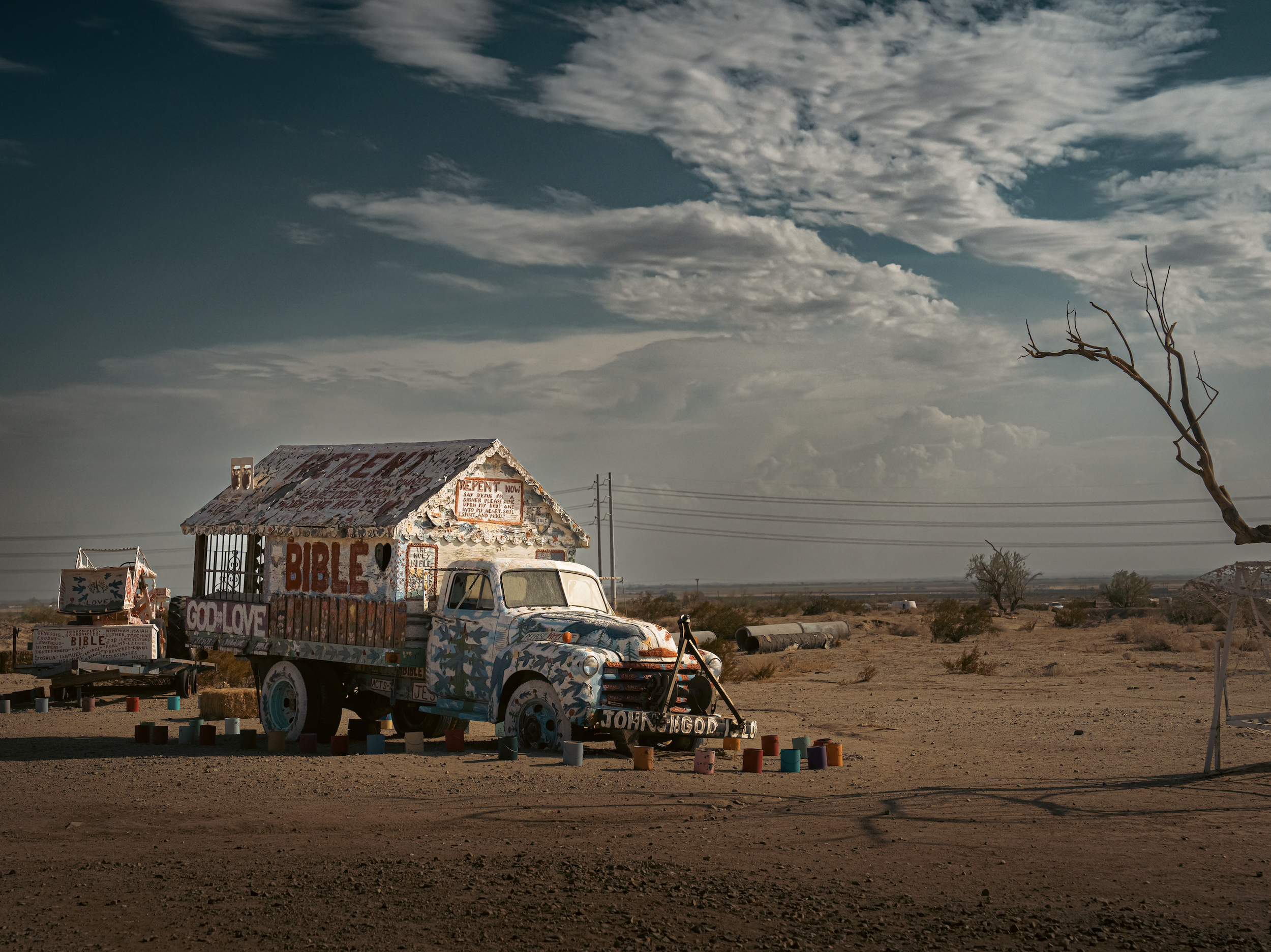 A small, colorful building decorated with religious signs, including a sign with the word 'BIBLE,' is situated in a desert landscape with a cloudy sky. An old, painted car is parked in front of the building, and a leafless tree is on the right side, with some scattered colorful objects on the ground.