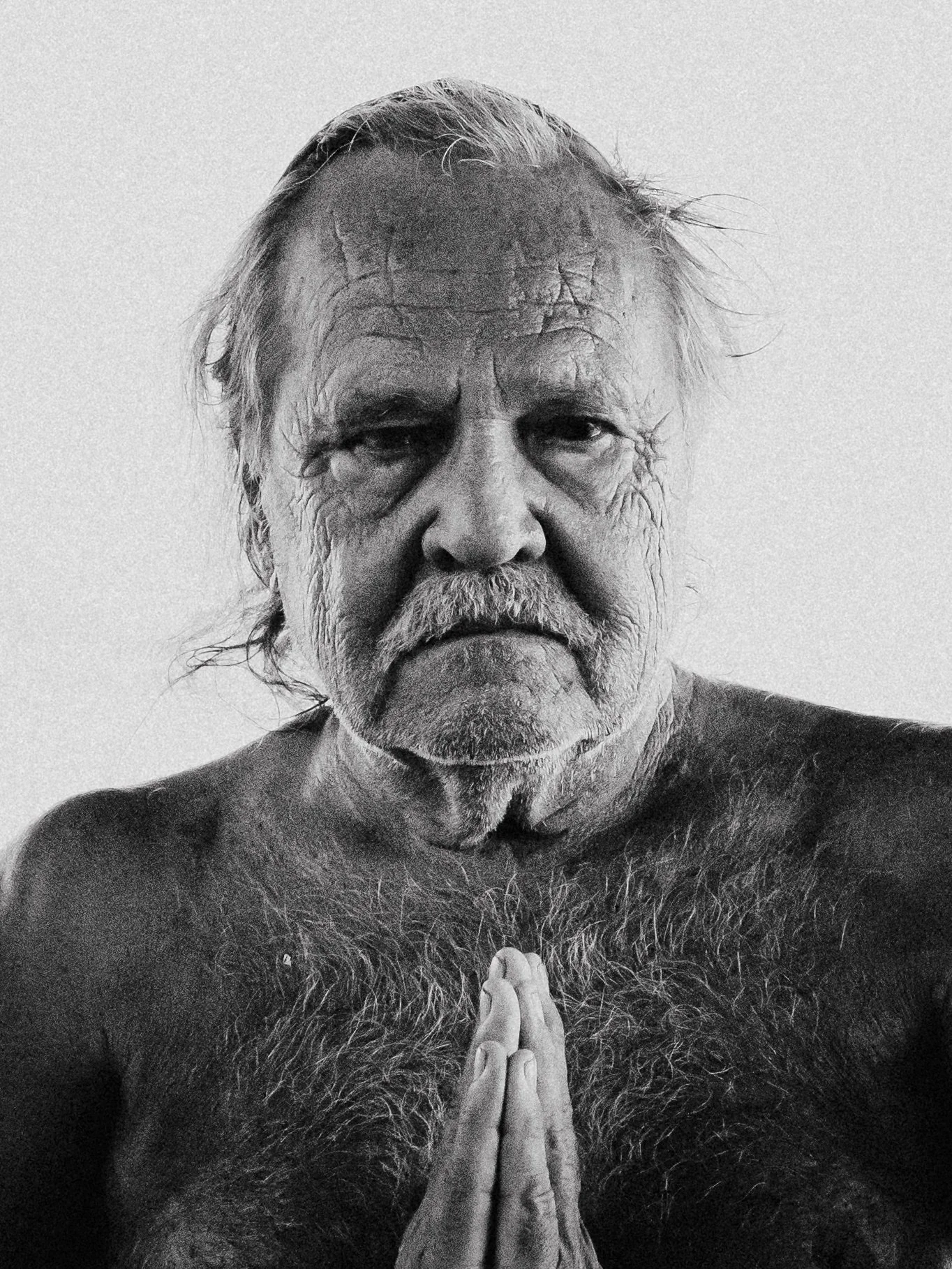 An elderly man with long gray hair and a beard, shirtless, pressing his palms together in a prayer position, standing against a plain background.
