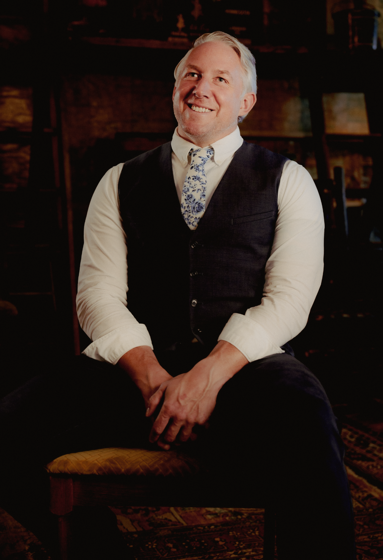 A smiling middle-aged man with gray hair, wearing a white shirt, dark vest, and floral tie, sitting on a chair with a patterned pillow, in a warmly lit room with wooden furniture in the background.