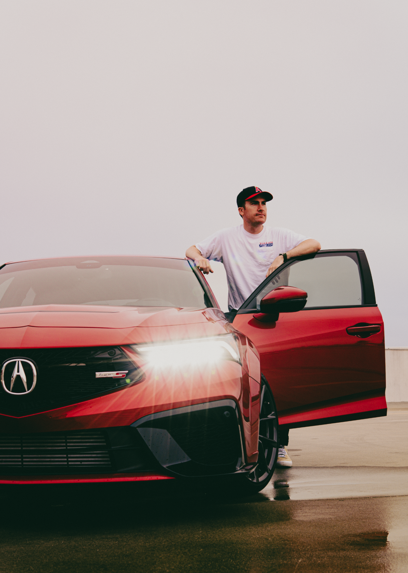 A man with dark hair wearing a white T-shirt and a black cap stands beside a red Acura car, resting his arm on the open driver's side door on a rooftop or parking lot with a plain sky in the background.