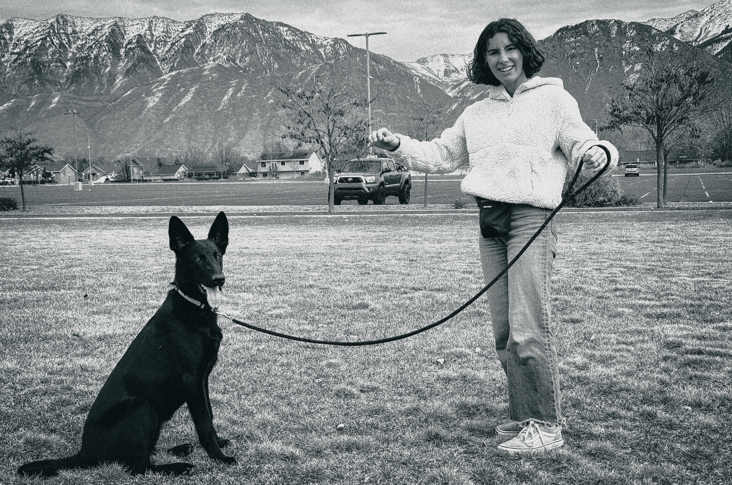 A woman with curly hair smiling while walking two dogs on leashes in a park with mountainous landscape in the background.