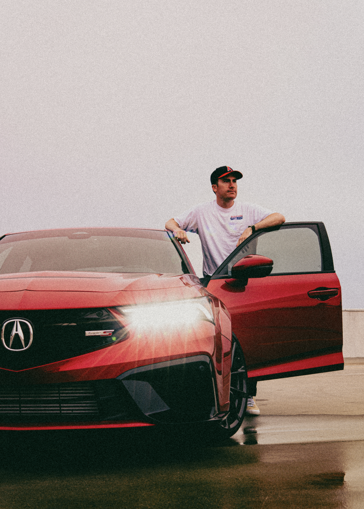 A man wearing a white t-shirt, black cap, and yellow shoes standing next to a red Acura car with the front and side visible, with a plain sky in the background.