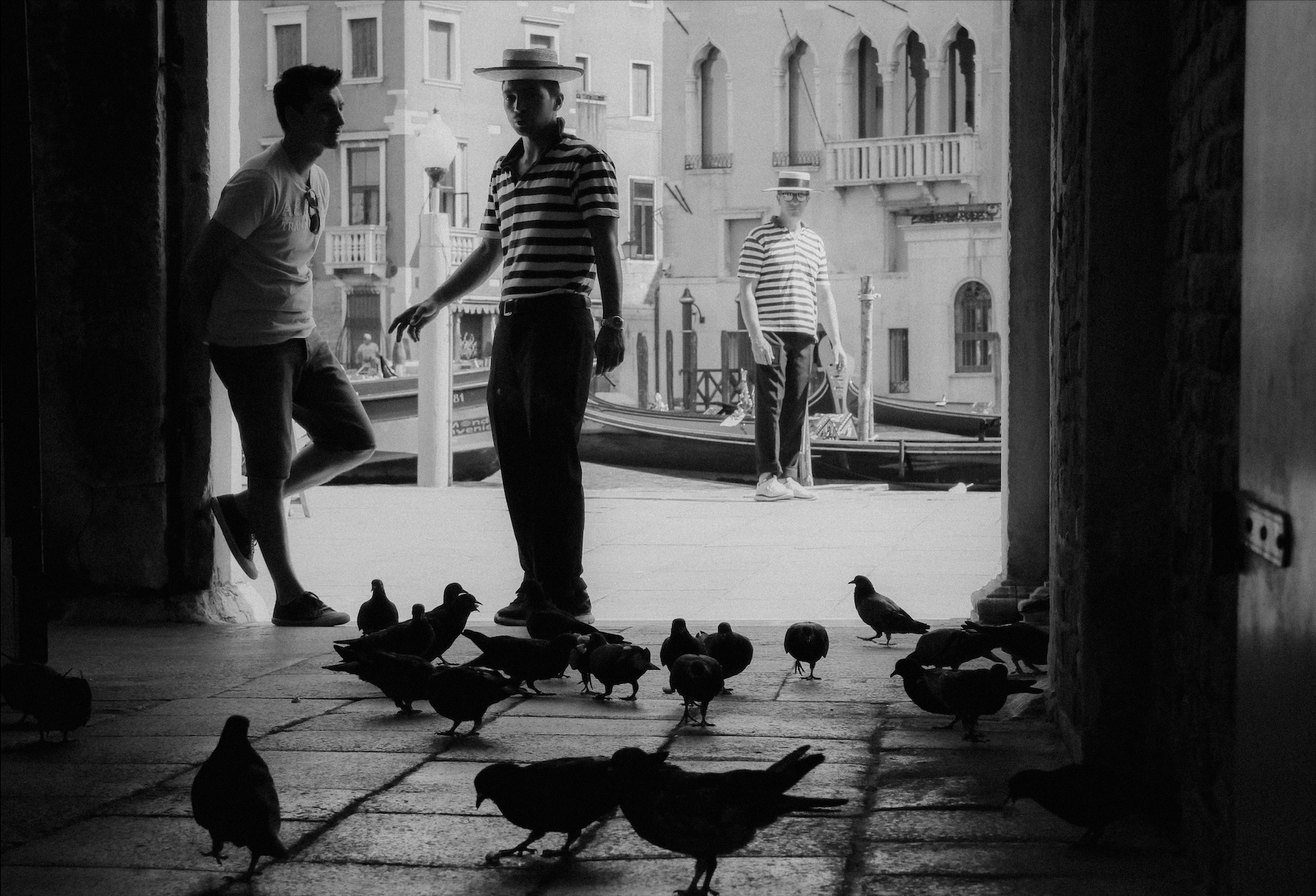 Black and white photo of three people standing near a canal with boats in the background. The person on the left is leaning against a stone pillar, the person in the center is wearing a striped shirt and a hat, and the person on the right is wearing 