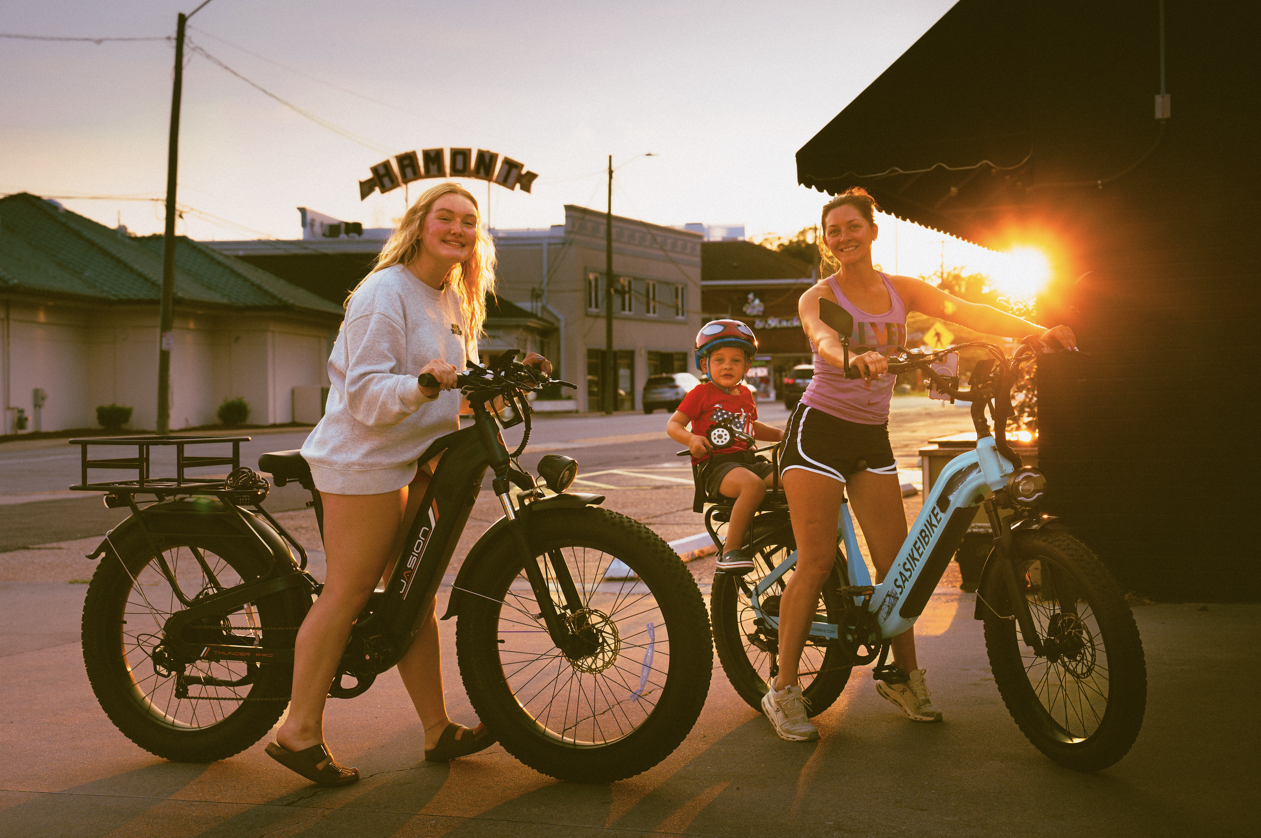 Three women and a young boy with a helmet on bicycles at sunset, standing on a street with buildings and a sign that reads 'HAMONT' behind them.