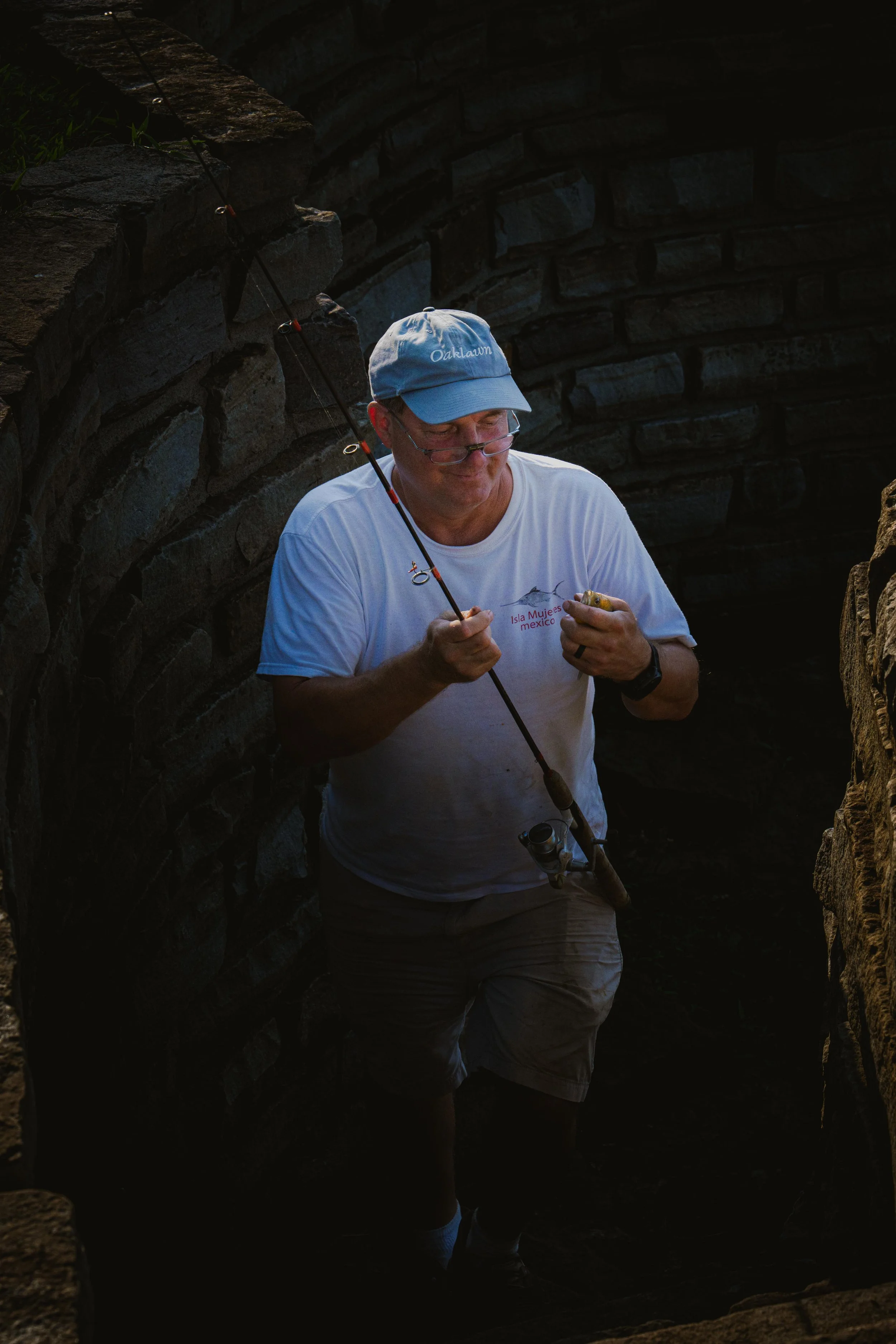 A man wearing a light-colored baseball cap, glasses, and a white t-shirt with red text and a fish design, is fishing with a fishing rod in a dark, rocky area.