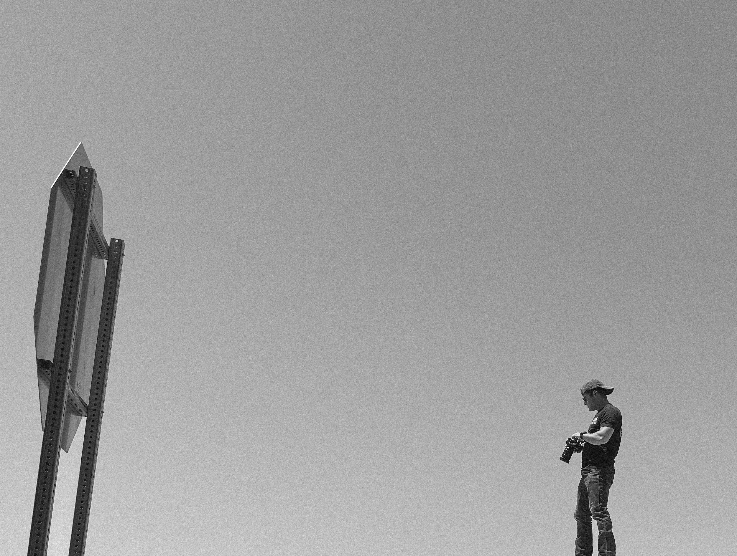 A man standing outdoors on a clear day, looking at his phone with a camera in his other hand, with a structure and sky in the background.