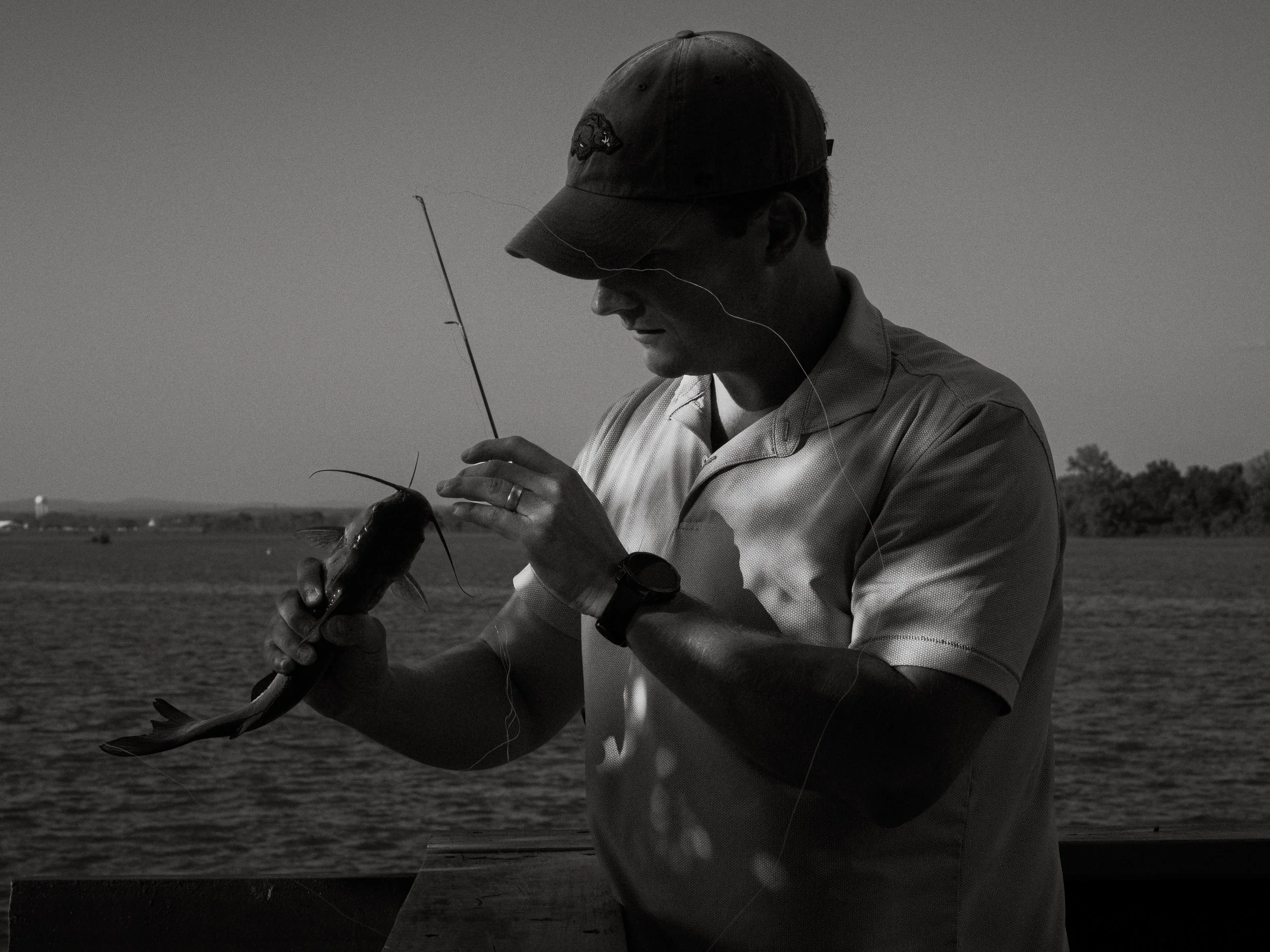 A man wearing a cap and shirt, holding a fish, standing outdoors near a body of water with a wide open landscape in the background, captured in black and white.