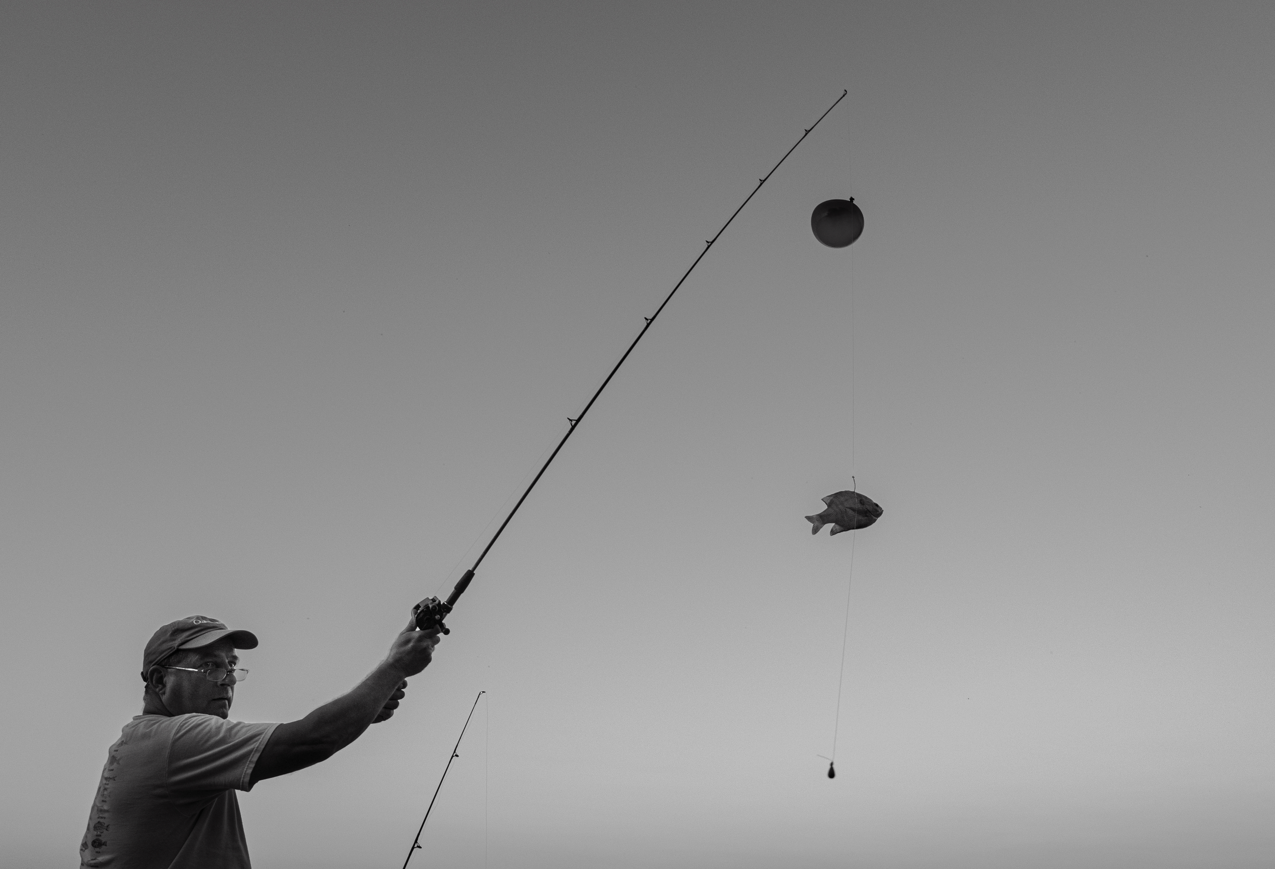 A man fishing with a fishing pole, a fish on the line, and a spherical bobber hanging from the line against a clear sky.