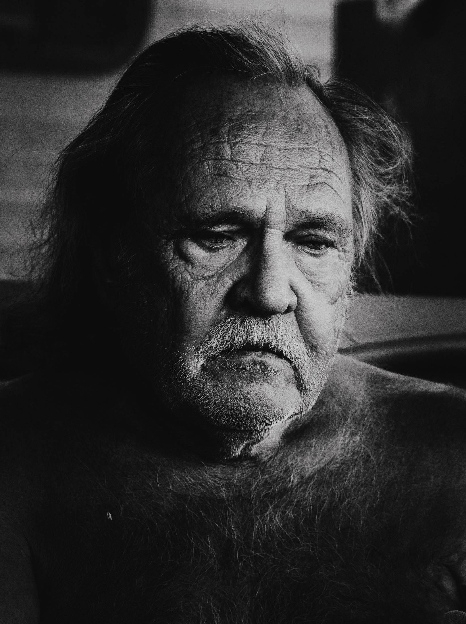 A close-up black and white photograph of an elderly man with a serious expression, displaying deep facial wrinkles, messy hair, and a white beard.