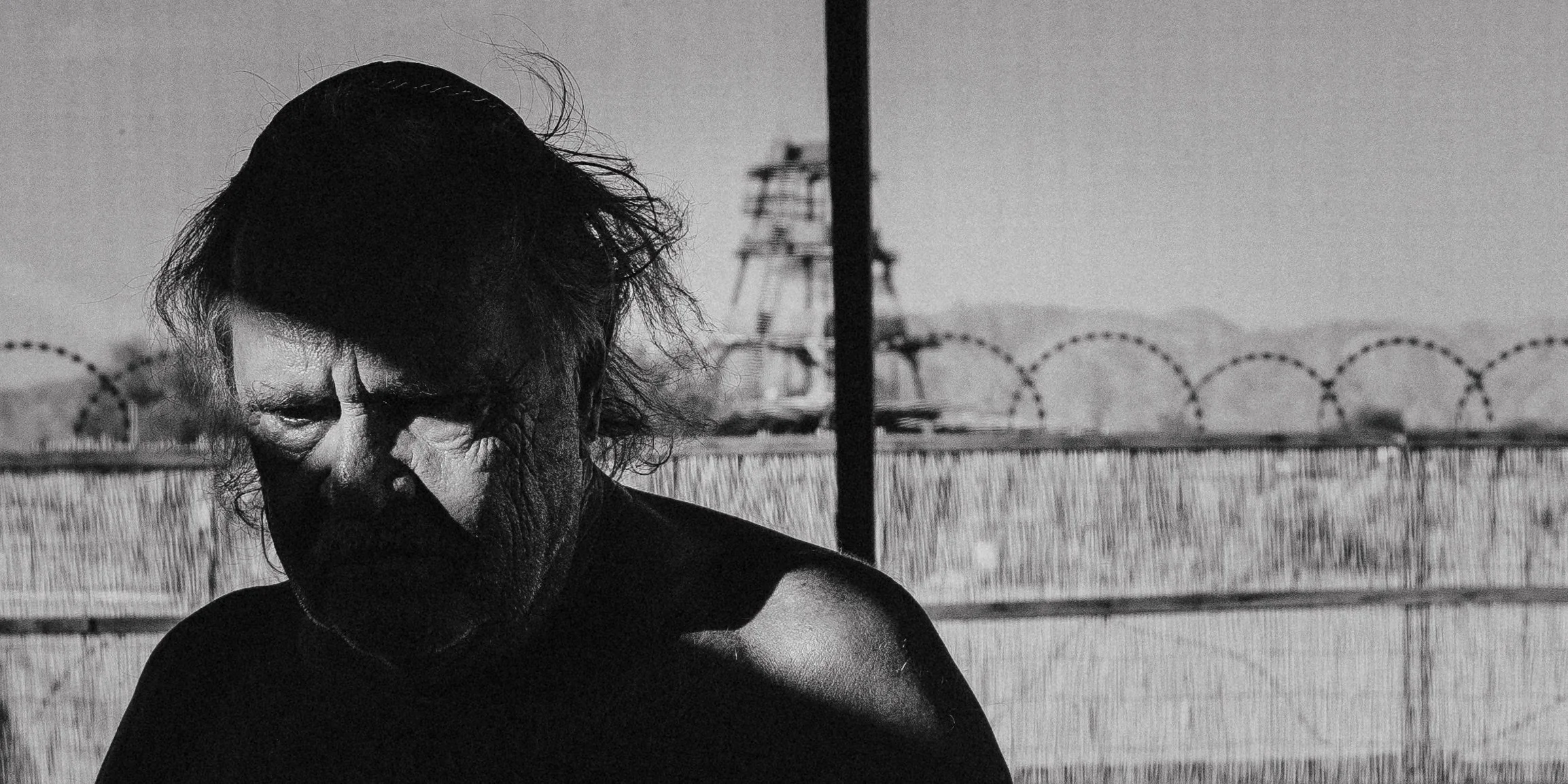 Black and white photo of a person's face, with disheveled hair and shadows obscuring parts of their face, standing in front of a fence and power lines in the background.