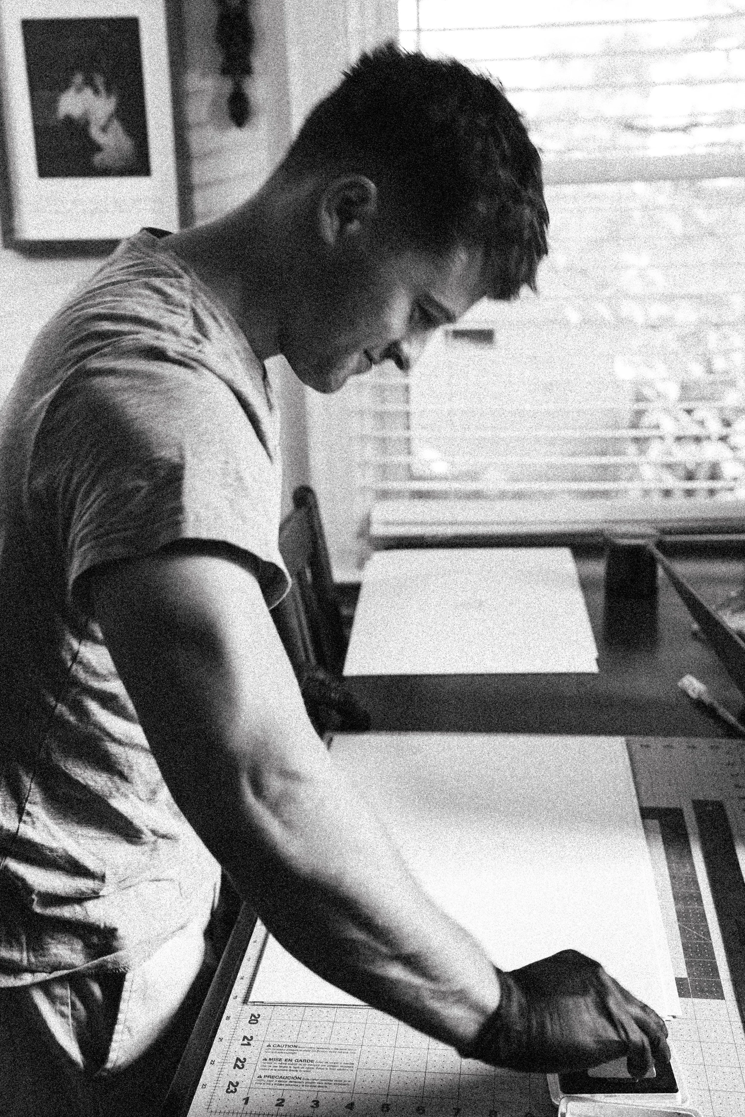 A person with short dark hair, wearing a T-shirt and gloves, is working on a cutting mat with a ruler and a pen or stylus, in a room with a window and a framed photograph of a dog on the wall.