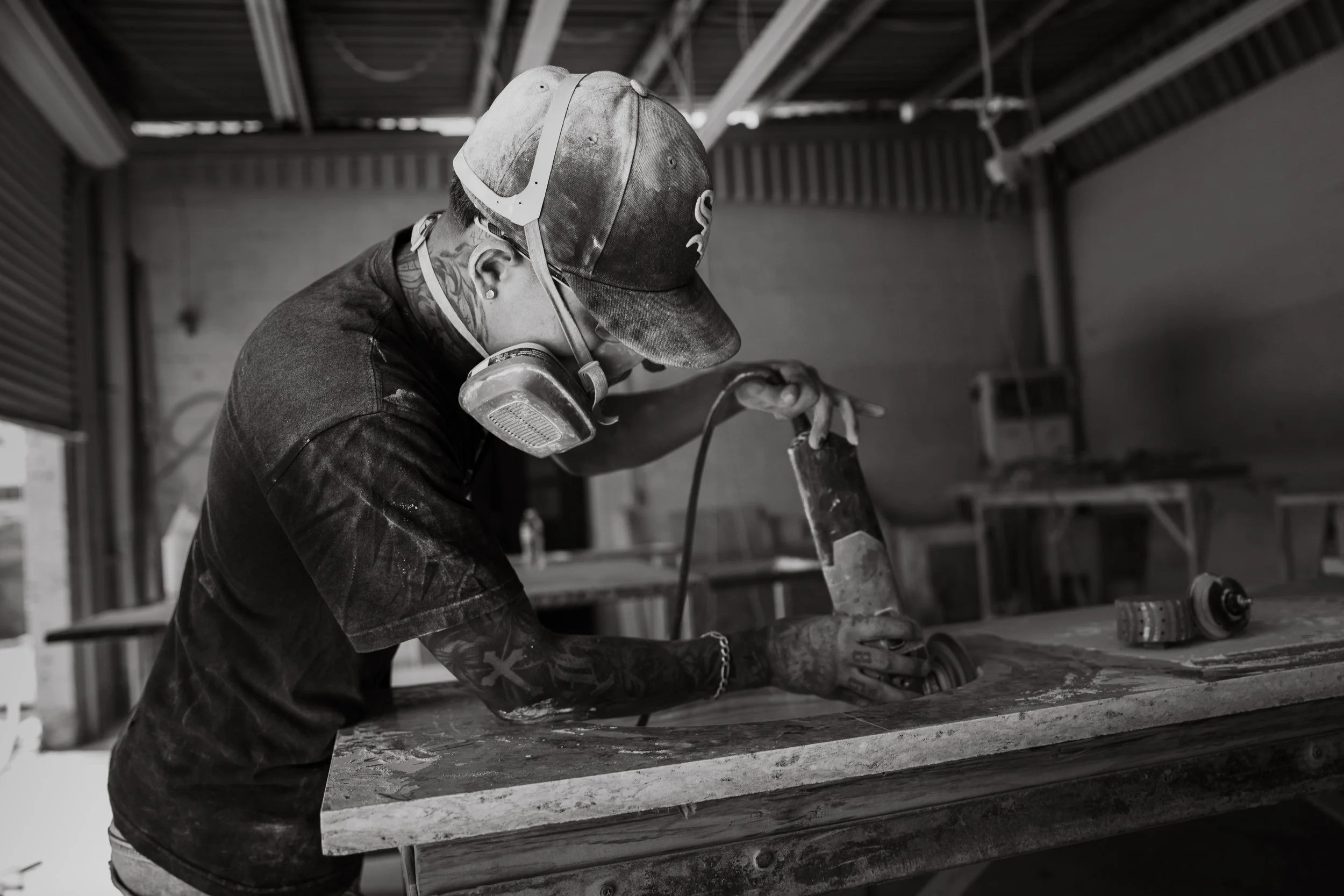 A person with tattoos and a cap wearing a mask is working on a project in a workshop, using a tool on a large piece of material.
