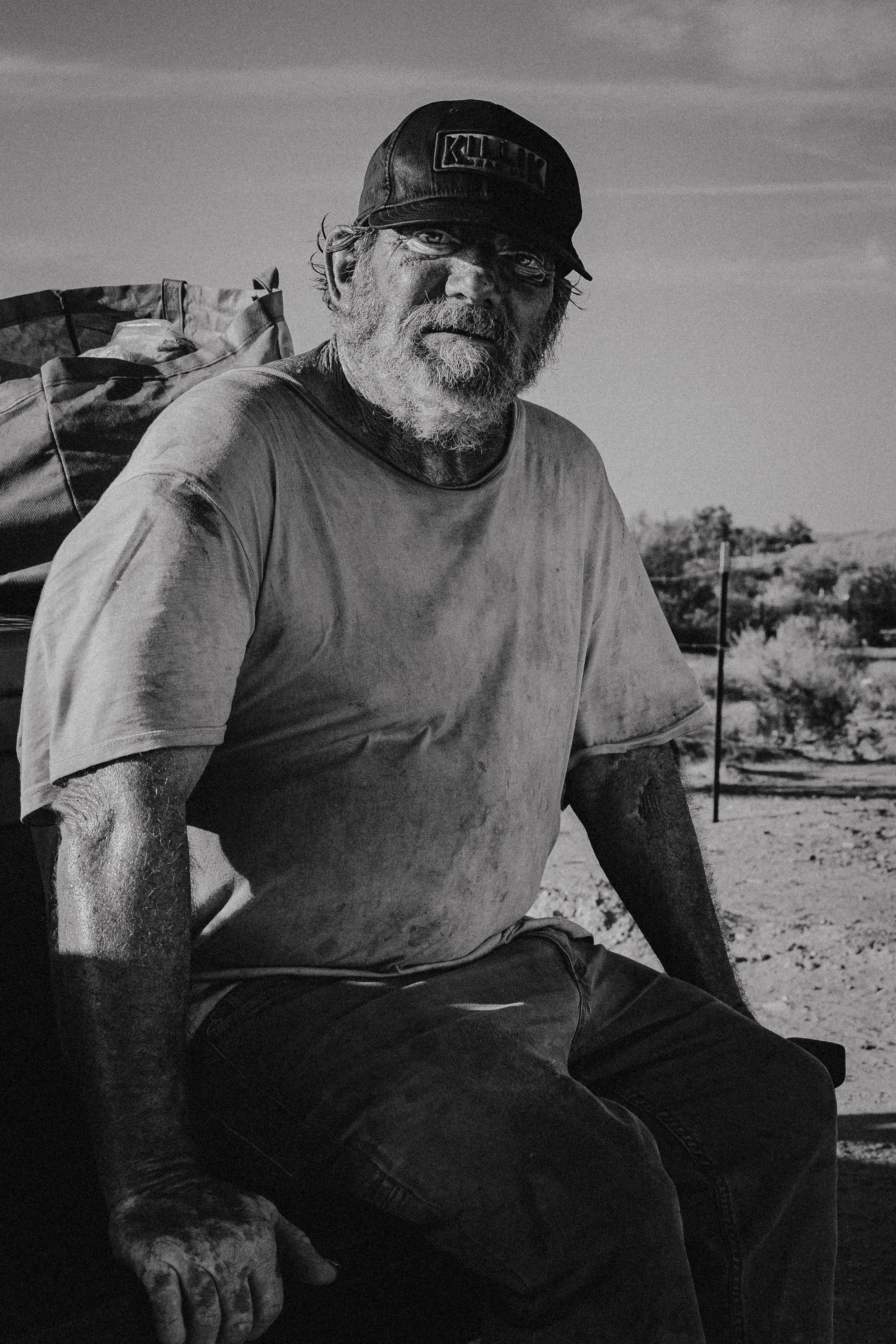 A black and white photo of an older man with a beard, wearing a cap labeled "RICH" and a t-shirt. He has a serious expression, with outdoor scenery characterized by dry terrain and sparse vegetation in the background.