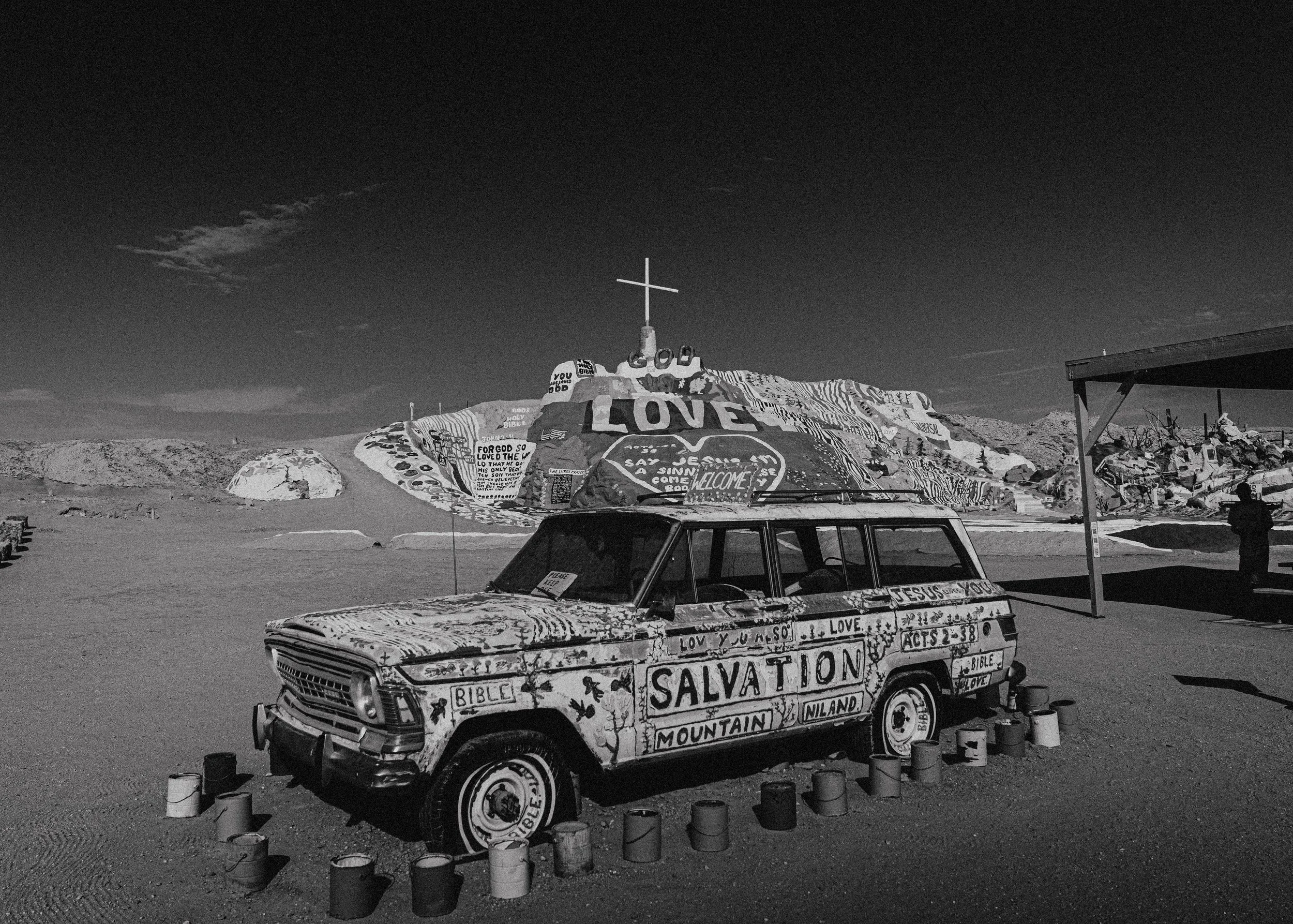 A painted vehicle in a desert landscape with hills and a cross on a hill in the background. The vehicle is covered in religious words and symbols, including 'LOVE,' 'SALVATION,' 'LOVE YOU SO,' and biblical references, along with various drawings and 