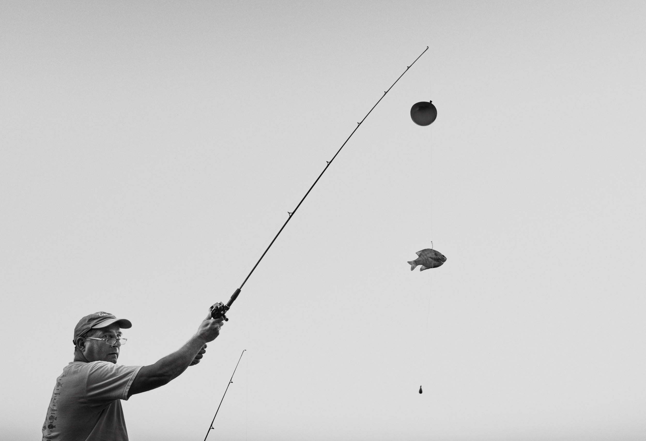 A man wearing glasses and a cap fishing with a rod, with a fish caught on the line, in black and white.