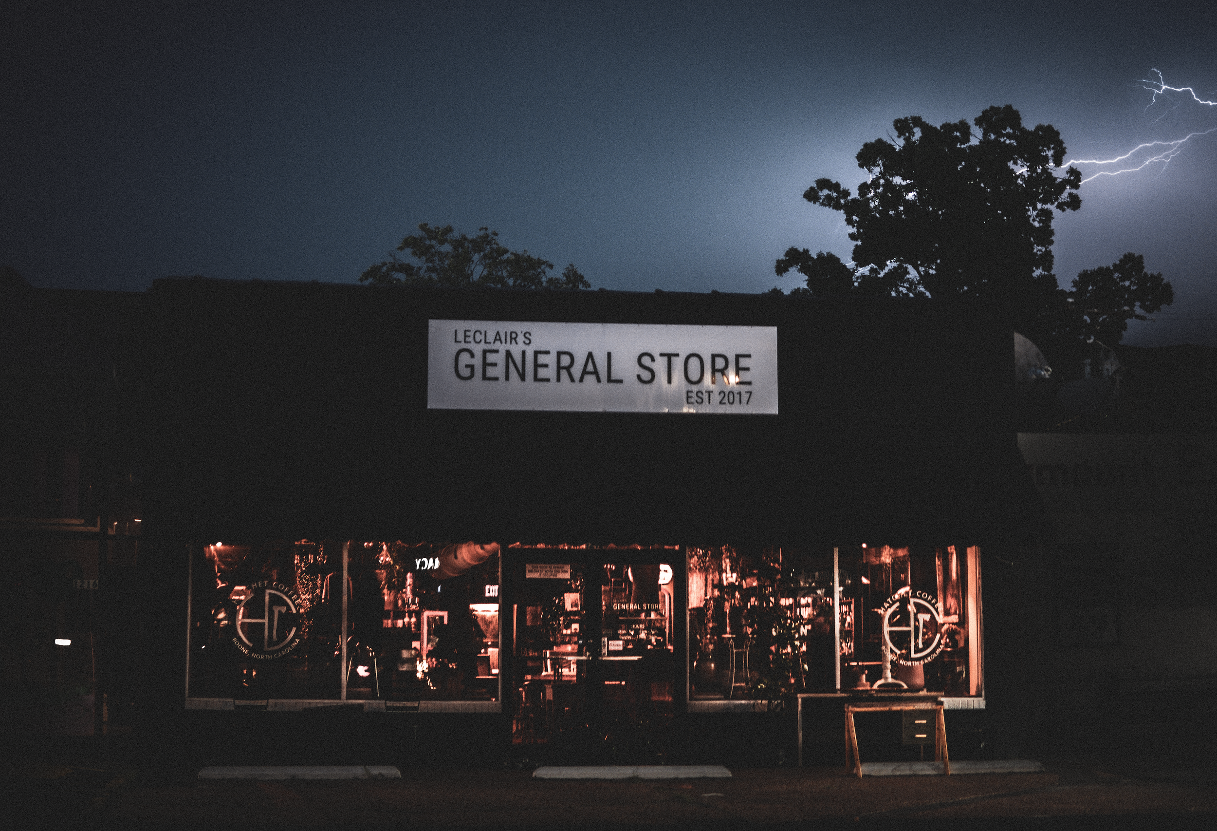 A store named 'Leclair's General Store' with a sign indicating it was established in 2017. The store is lit from inside, and there are trees and lightning in the dark sky above.