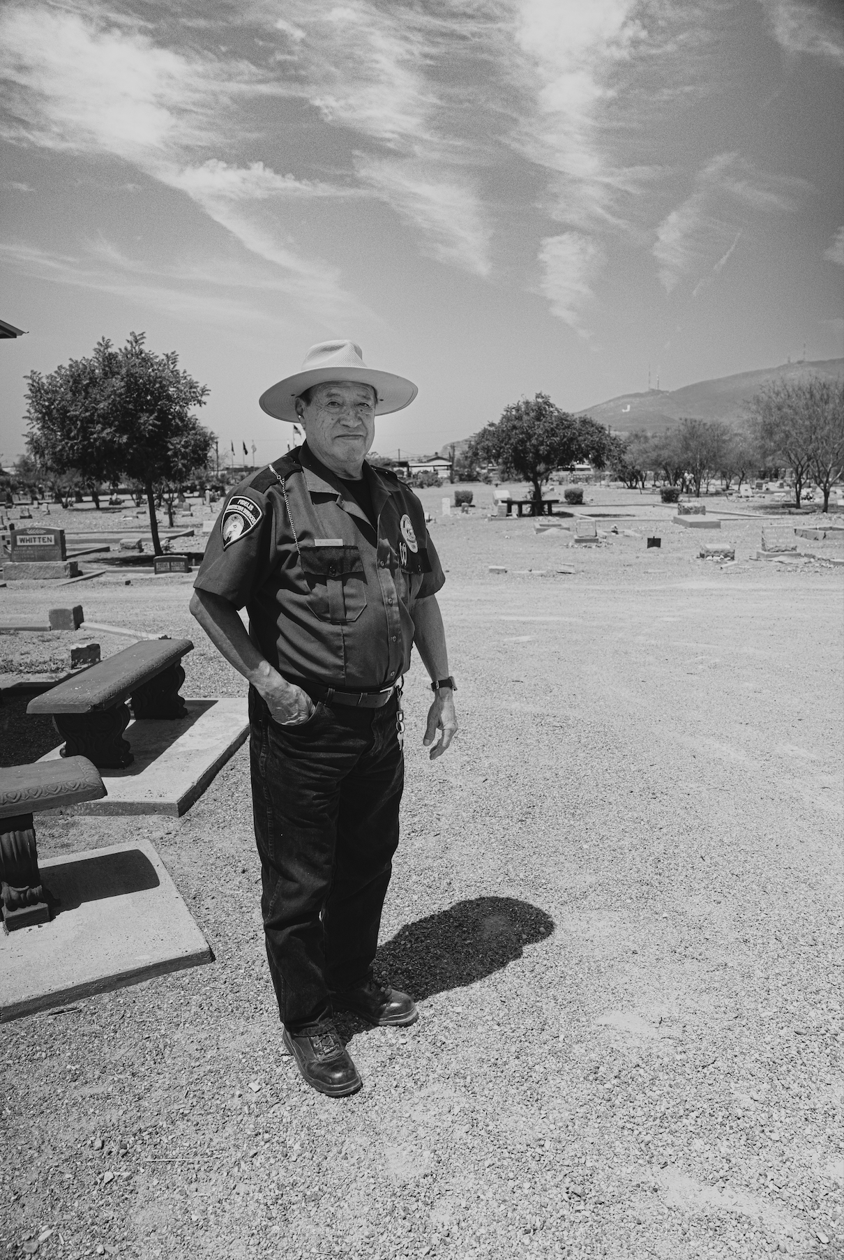 A man in a police or security uniform and a wide-brimmed hat standing outdoors in a cemetery. The background features scattered trees, tombstones, and a hill with communication towers.