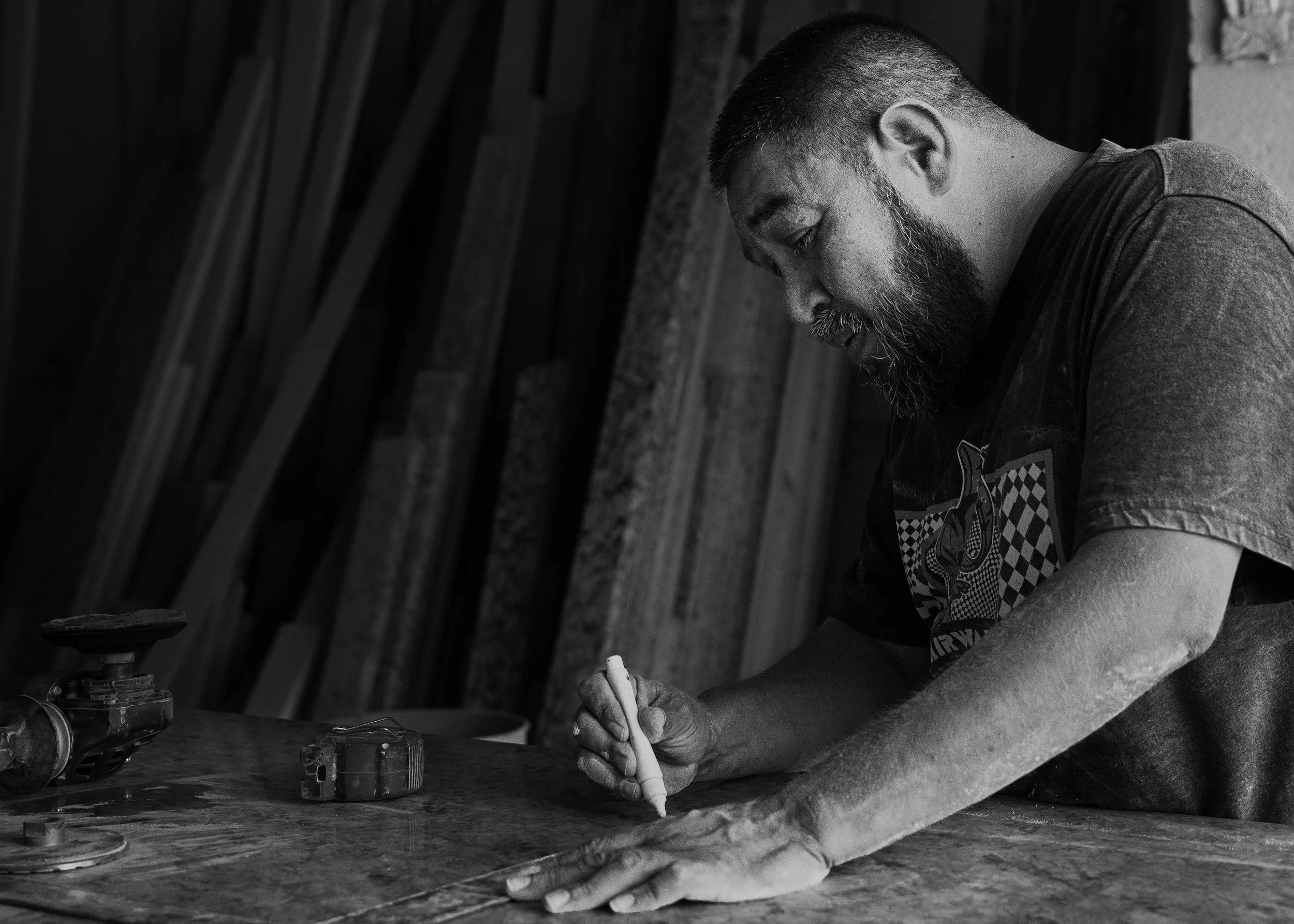 A man with a beard and short hair is writing on a piece of paper at a workbench in a workshop. The workshop has wooden beams in the background, and various tools and equipment are placed on the work surface.