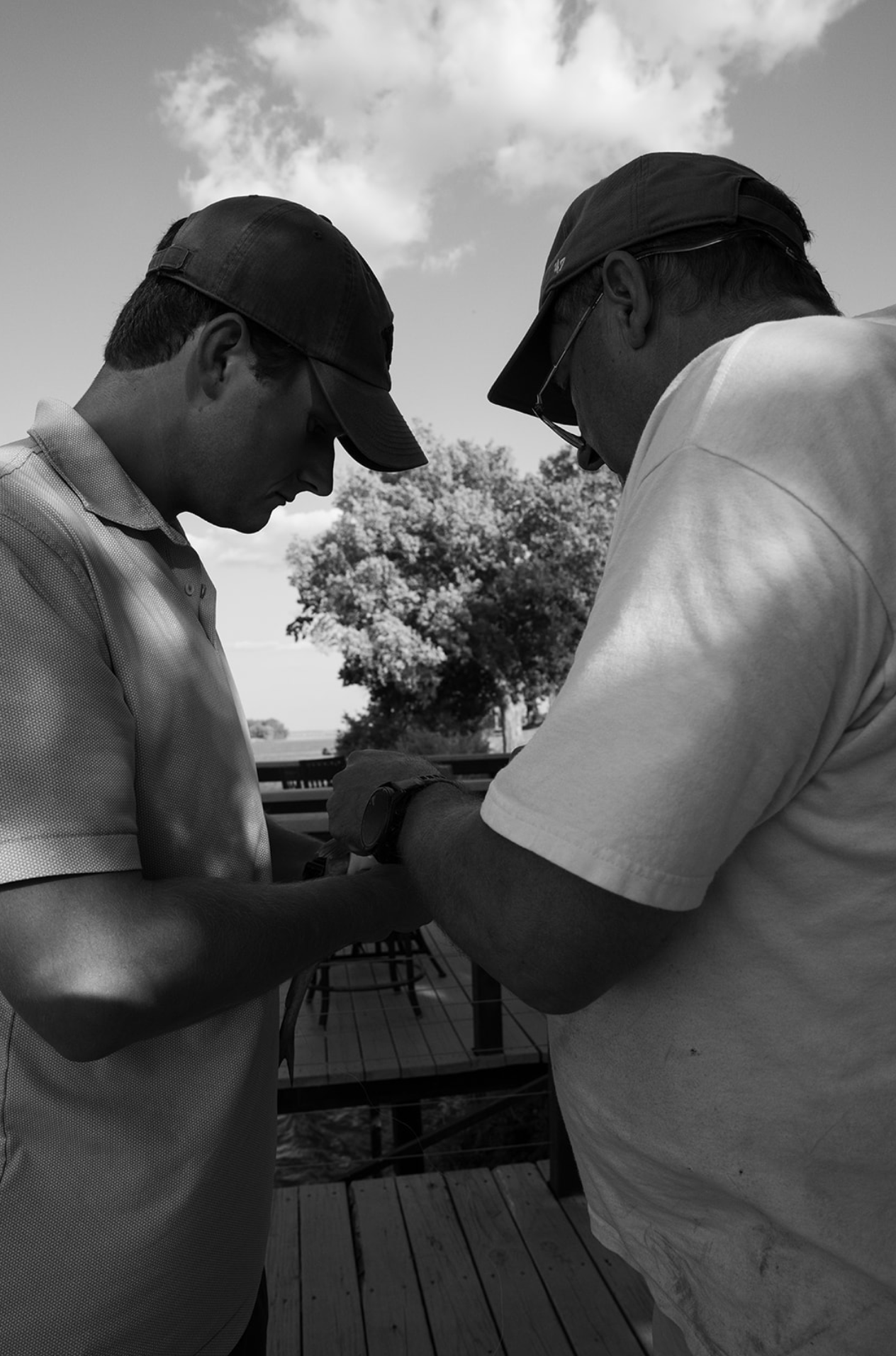 Two men wearing caps and casual clothing look down at an object, engaging in a focused activity outdoors on a wooden deck, with trees and clouds in the background.
