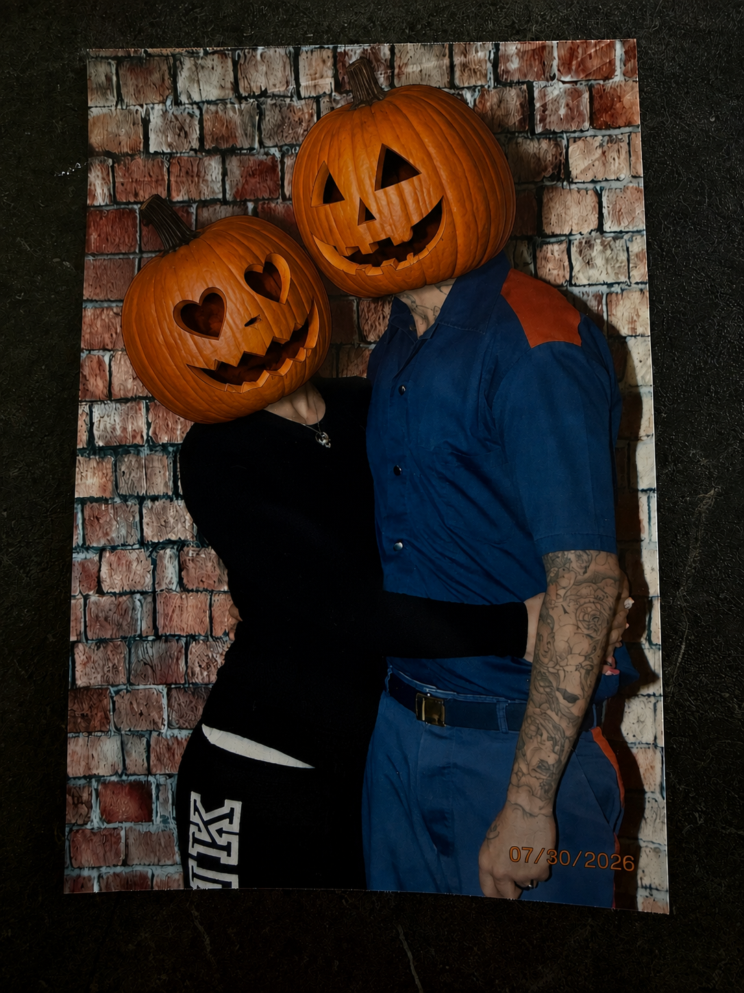 Two people with pumpkin heads, carved as Jack-o'-lanterns, posing in front of a brick wall backdrop. One person's head has heart-shaped eyes, while the other's has triangle eyes and a big smile. One person is wearing a black outfit, and the other is dressed in a blue shirt with rolled-up sleeves and tattoos on their arm.