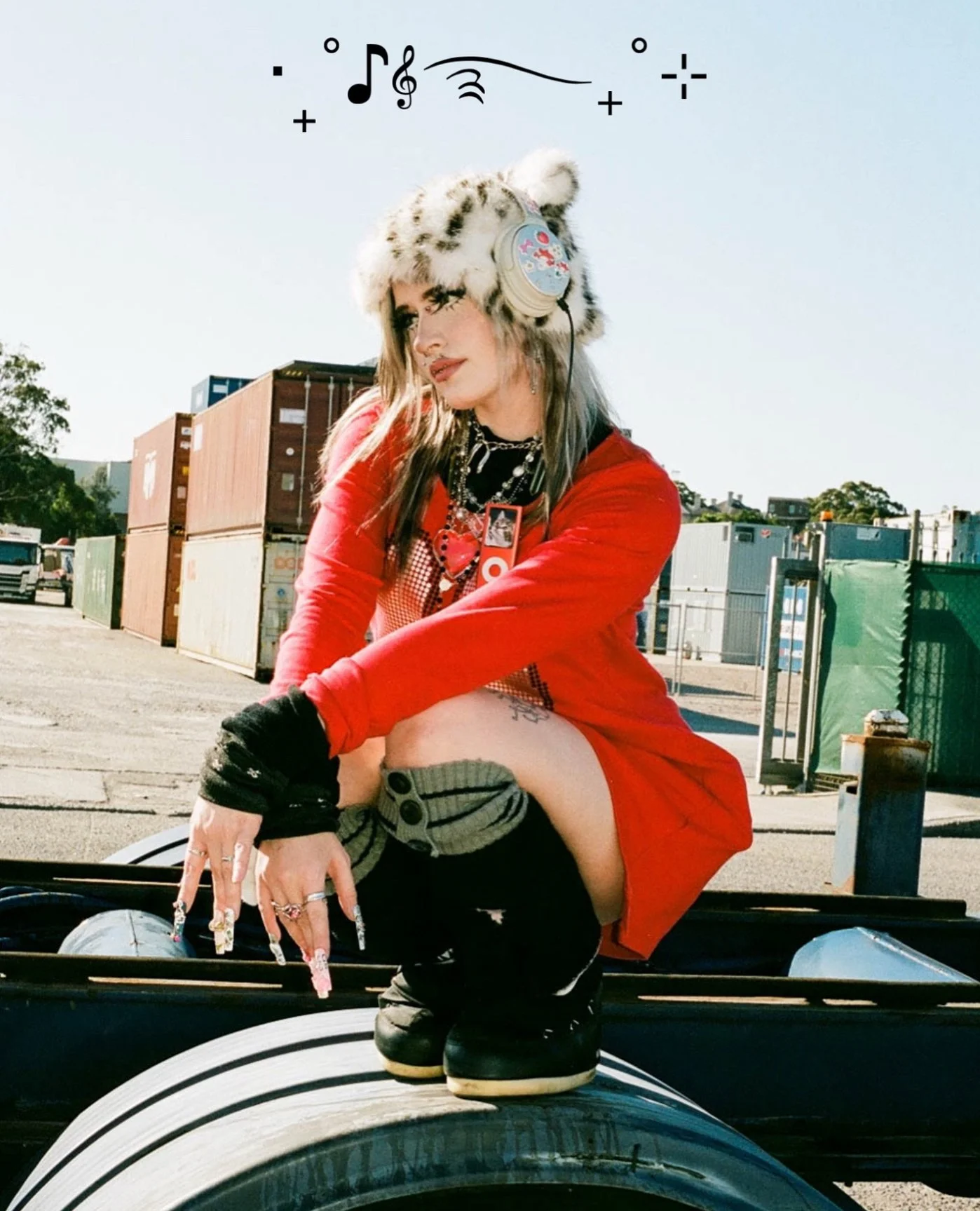 Young woman with colorful hair, wearing a furry hat, headphones, and a red jacket, sitting on a vehicle in an industrial outdoor setting with containers in the background.