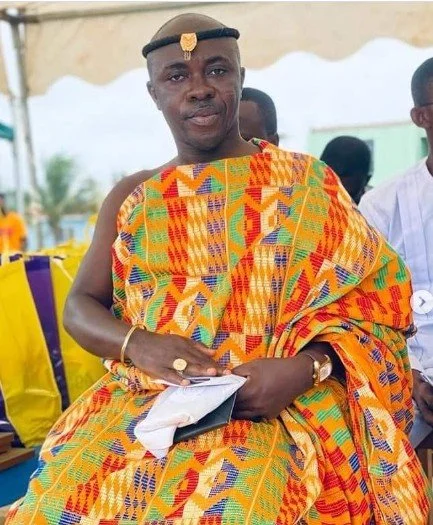 A man dressed in traditional colorful kente cloth sitting on a chair at an outdoor event, wearing a cap and holding a piece of paper.