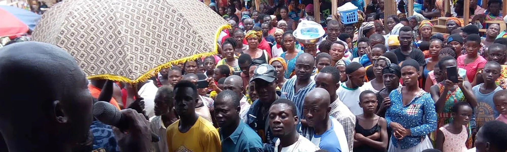 A large crowd of people gathered outdoors, with a woman speaking into a microphone and holding an umbrella under the sun.