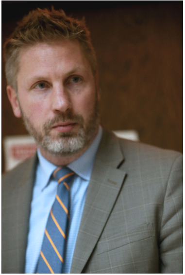 A man with light brown hair and a beard wearing a light gray suit and a blue shirt with a striped tie, looking to the side.
