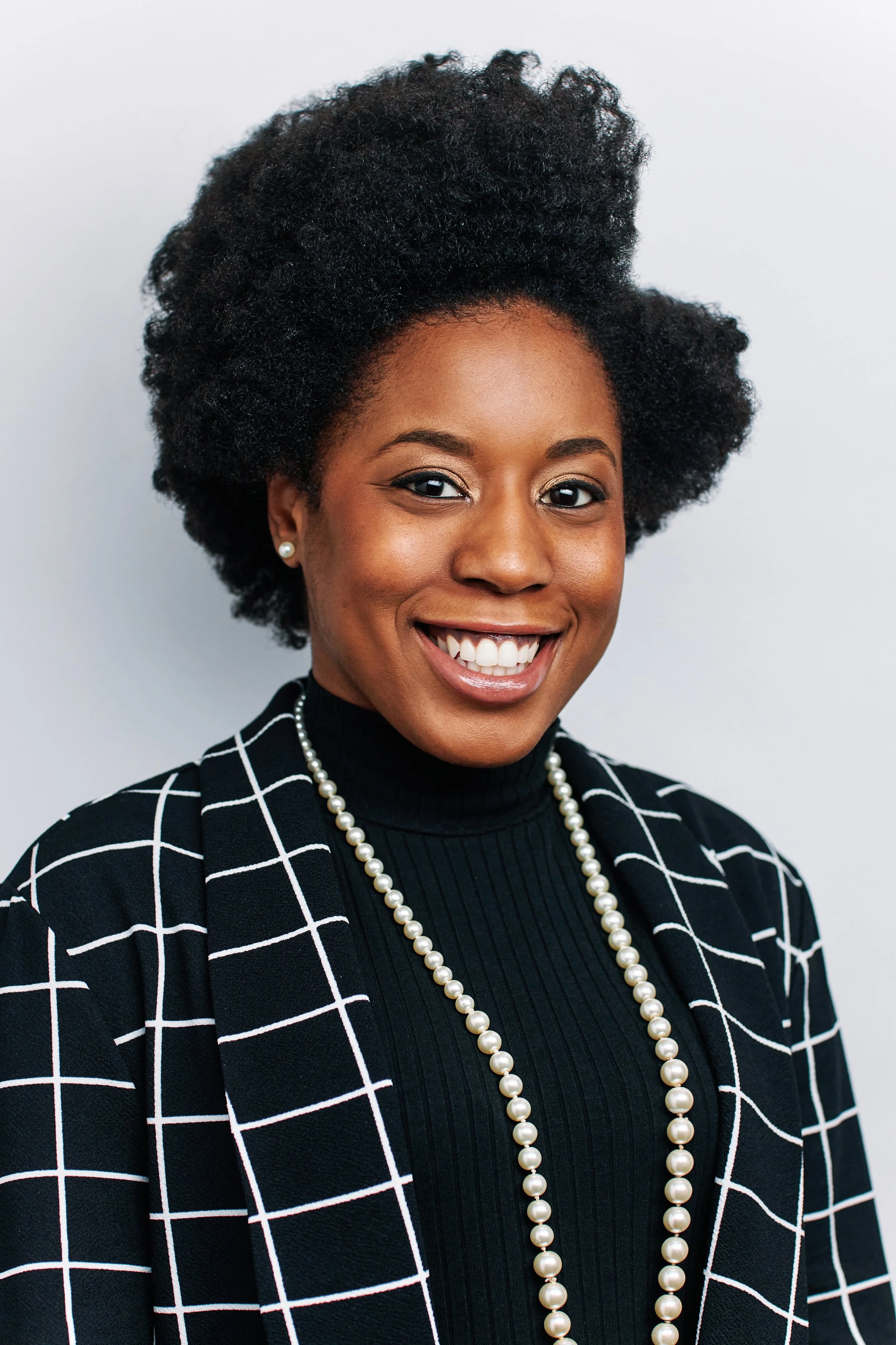 A smiling woman wearing a black turtleneck, a black and white checkered blazer, pearl earrings, and a pearl necklace. She has natural curly black hair styled voluminously.