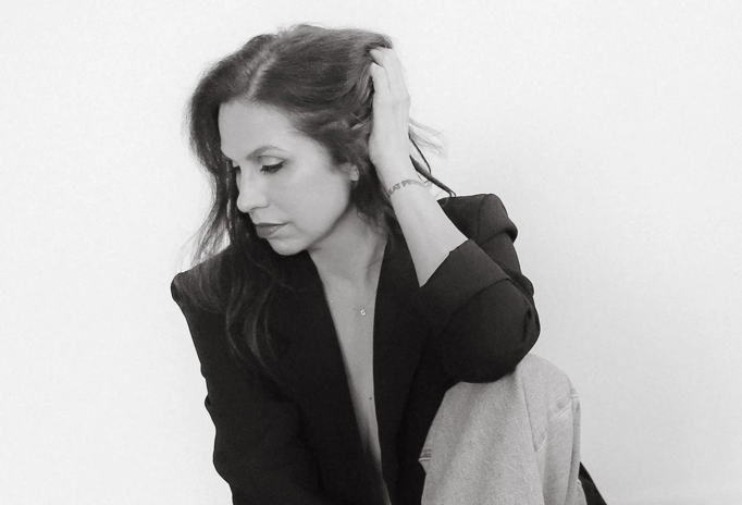 Black and white photo of a woman with long hair, wearing a blazer, sitting against a plain background, looking down with her hand on the back of her head.