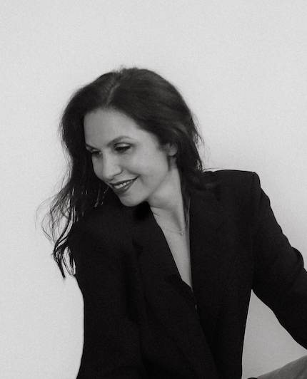 A well-dressed therapist with long dark hair smiling and looking down, wearing a black blazer, against a plain background.