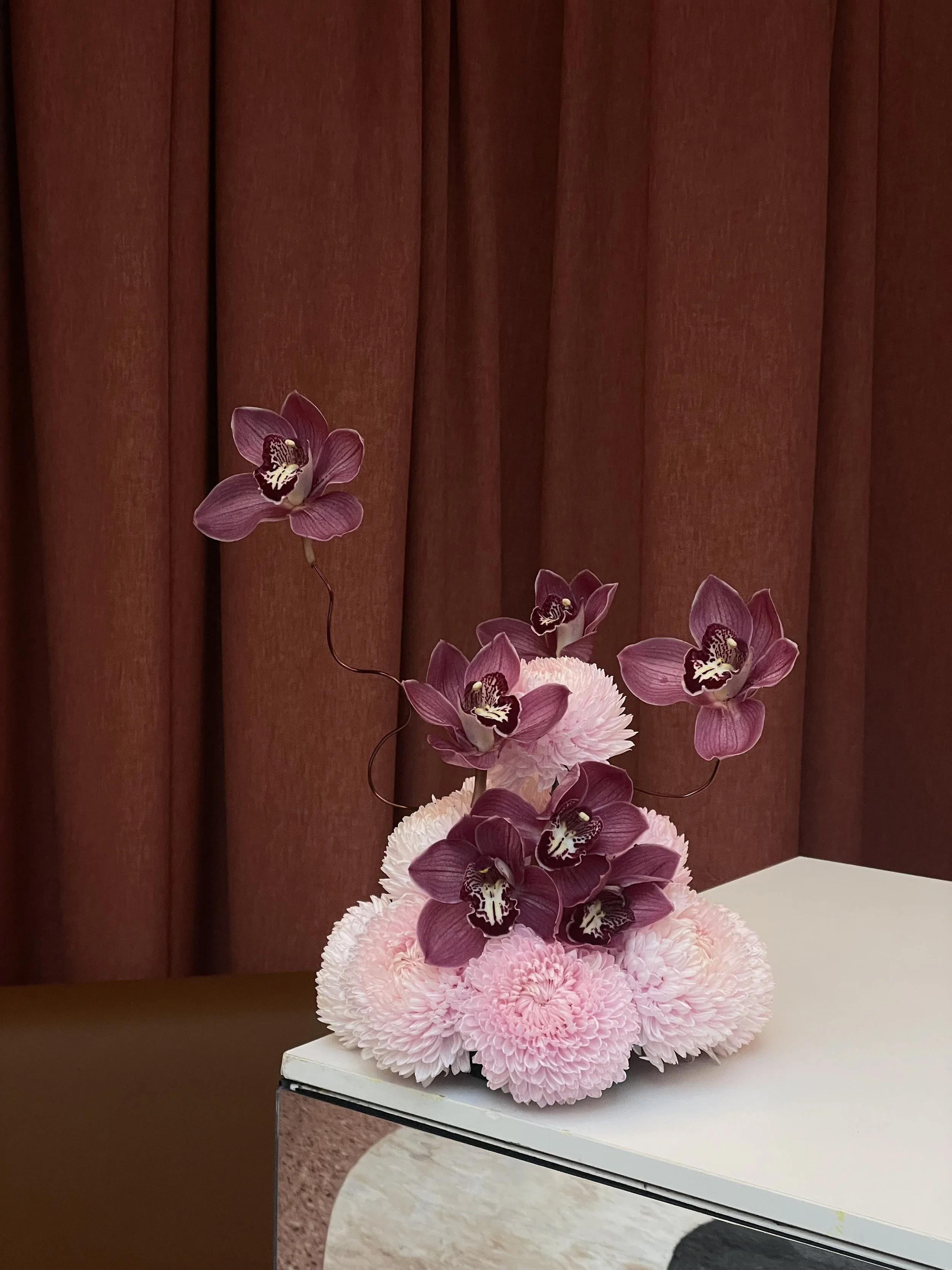 A floral arrangement featuring pink carnations and purple orchids on a white surface against a brown curtain background.