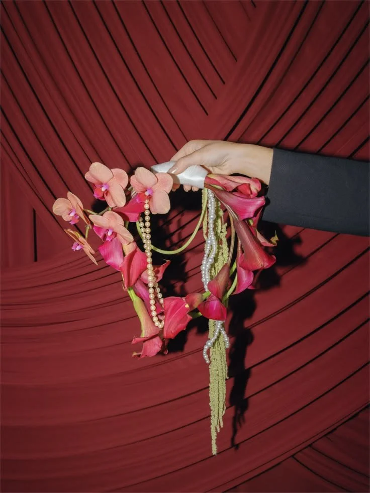 A hand holding a heart-shaped floral arrangement made of pink orchids and pink calla lilies, adorned with pearl necklaces, with a red, pleated fabric background.