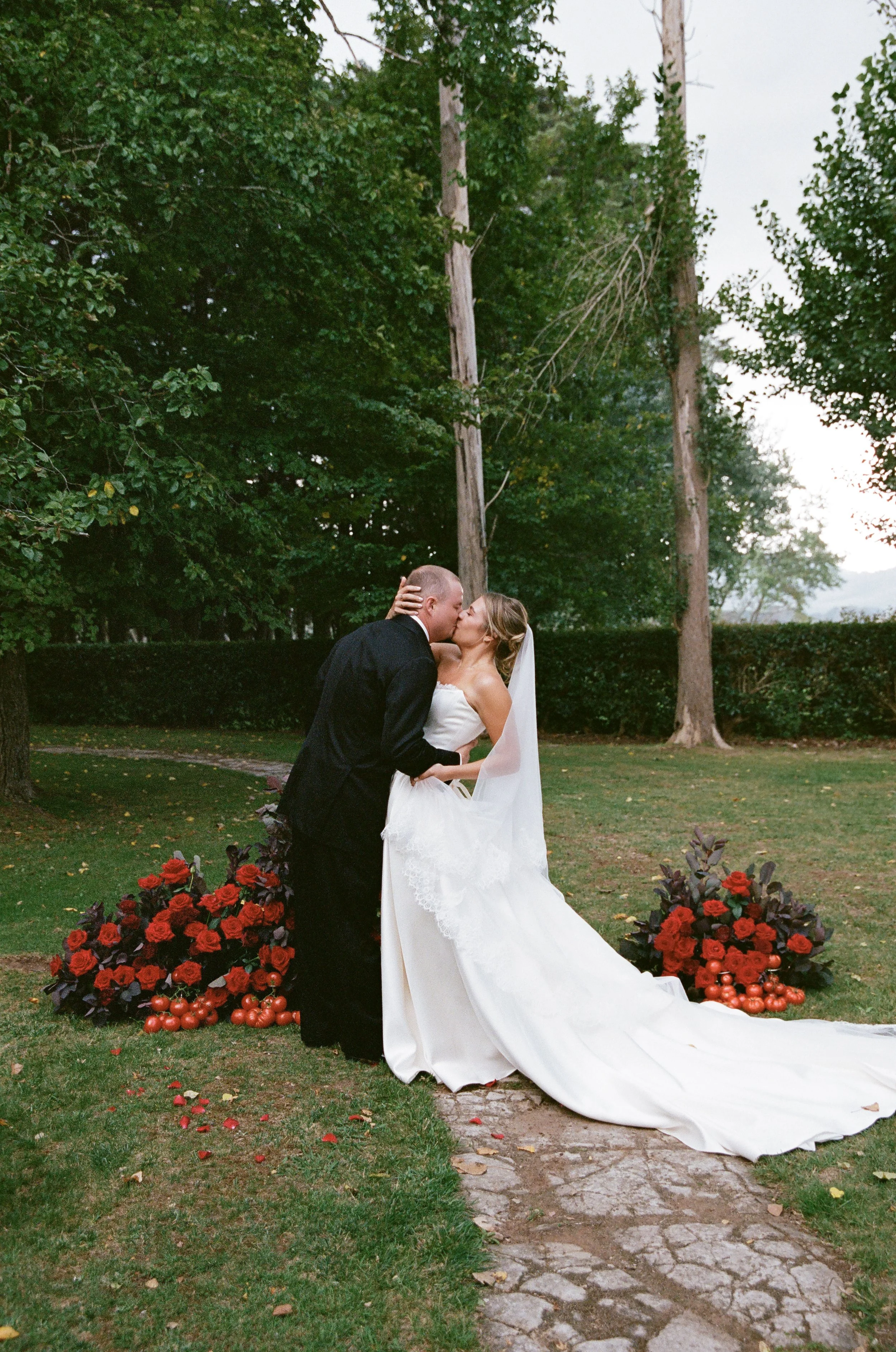 A bride and groom share a kiss outdoors during their wedding ceremony. The bride wears a white wedding gown with a train, and the groom wears a black suit. There are two large flower arrangements with red and pink flowers at their sides, and a red rose petal path on the ground. The background features green trees and a cloudy sky.