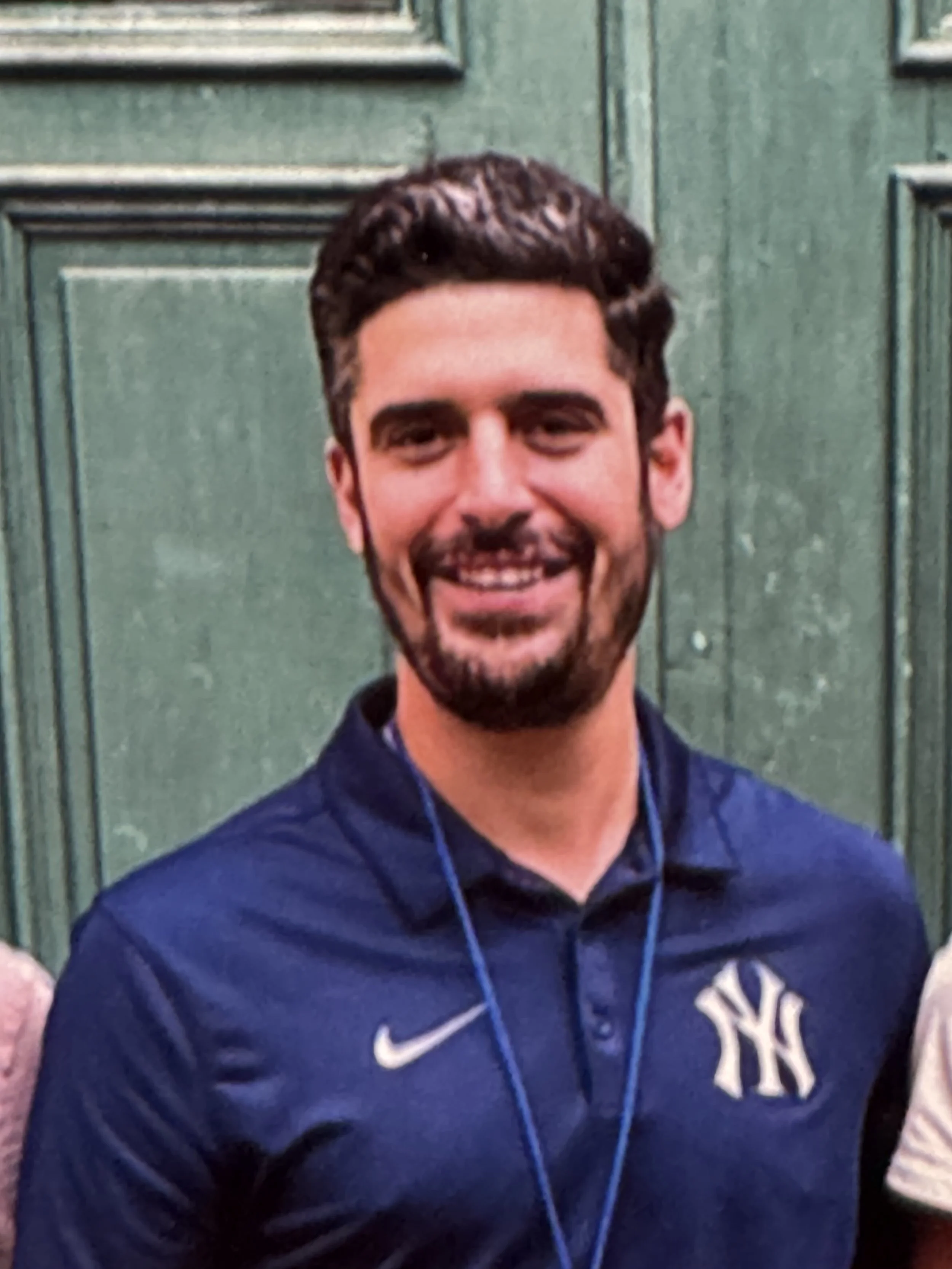A young man with dark hair and a beard, smiling, wearing a navy blue sports jacket with a white NY logo, standing in front of green wooden panels.