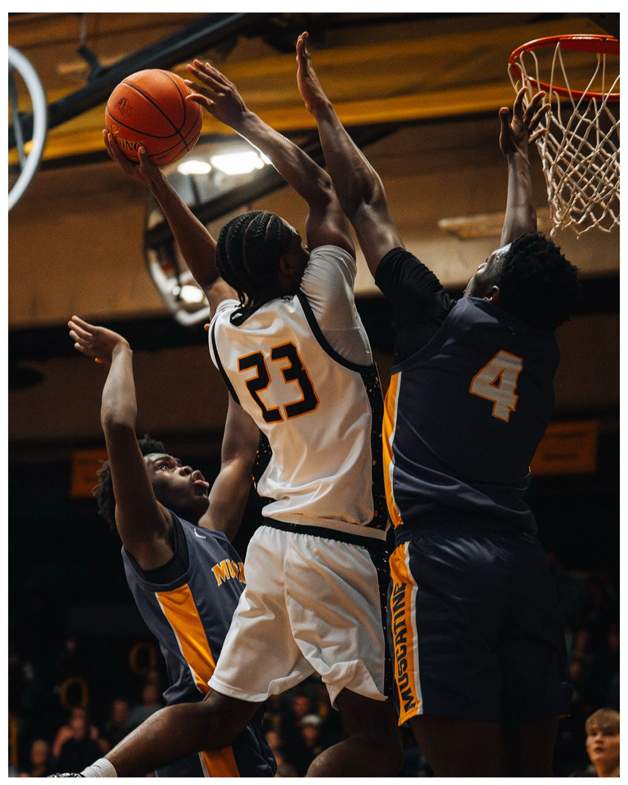 Two basketball players mid-game competing for the ball near the basket. One player is wearing a white jersey with the number 23, and the other is in a dark jersey with the number 4. The scene is indoors with a basketball hoop and net visible in the background.
