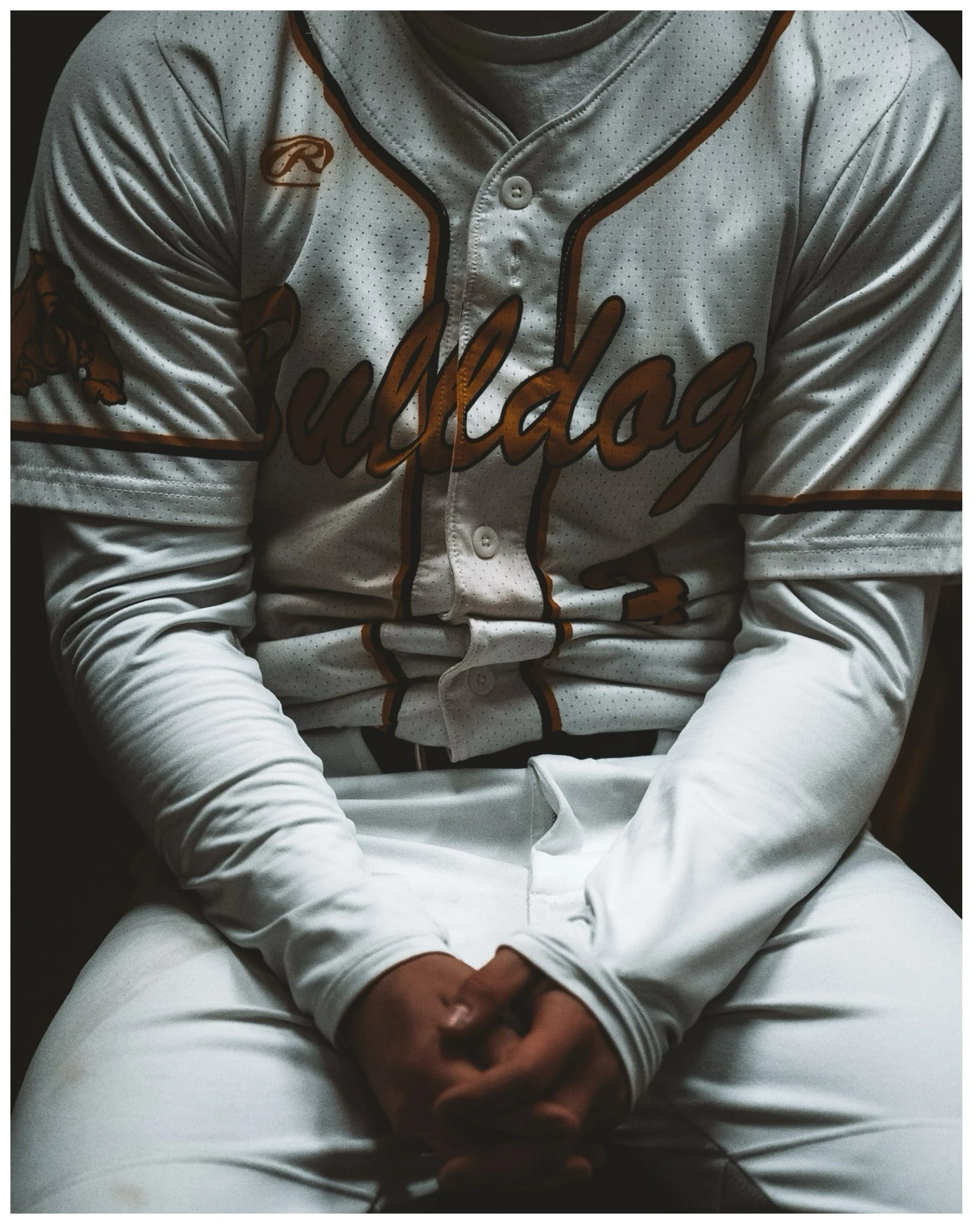 Person wearing a white baseball jersey with orange and black accents, sitting with hands clasped in front of them, the jersey has the text 'Baylor' on it and a Rawlings logo.
